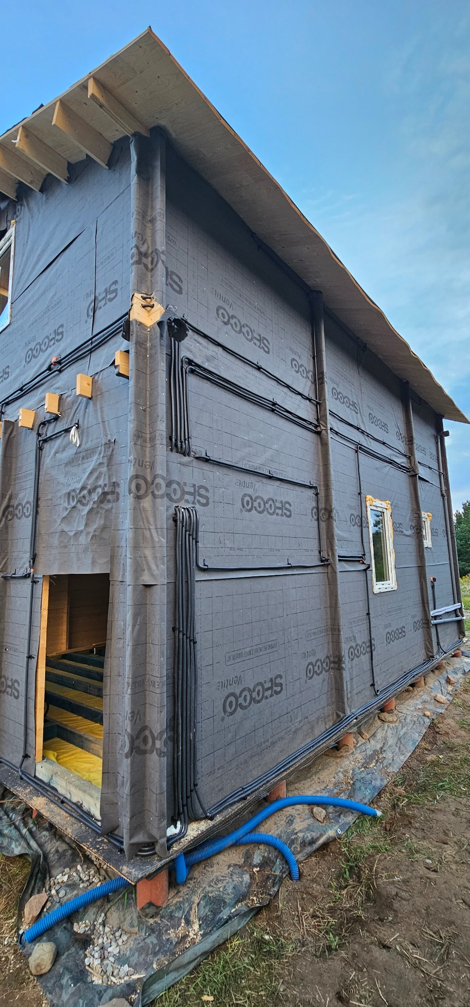 Dom w trakcie budowy z widoczną instalacją elektryczną na zewnątrz. Czarne rury prowadzone po ścianach pokrytych membraną. Widoczne okna i fragment konstrukcji dachu.