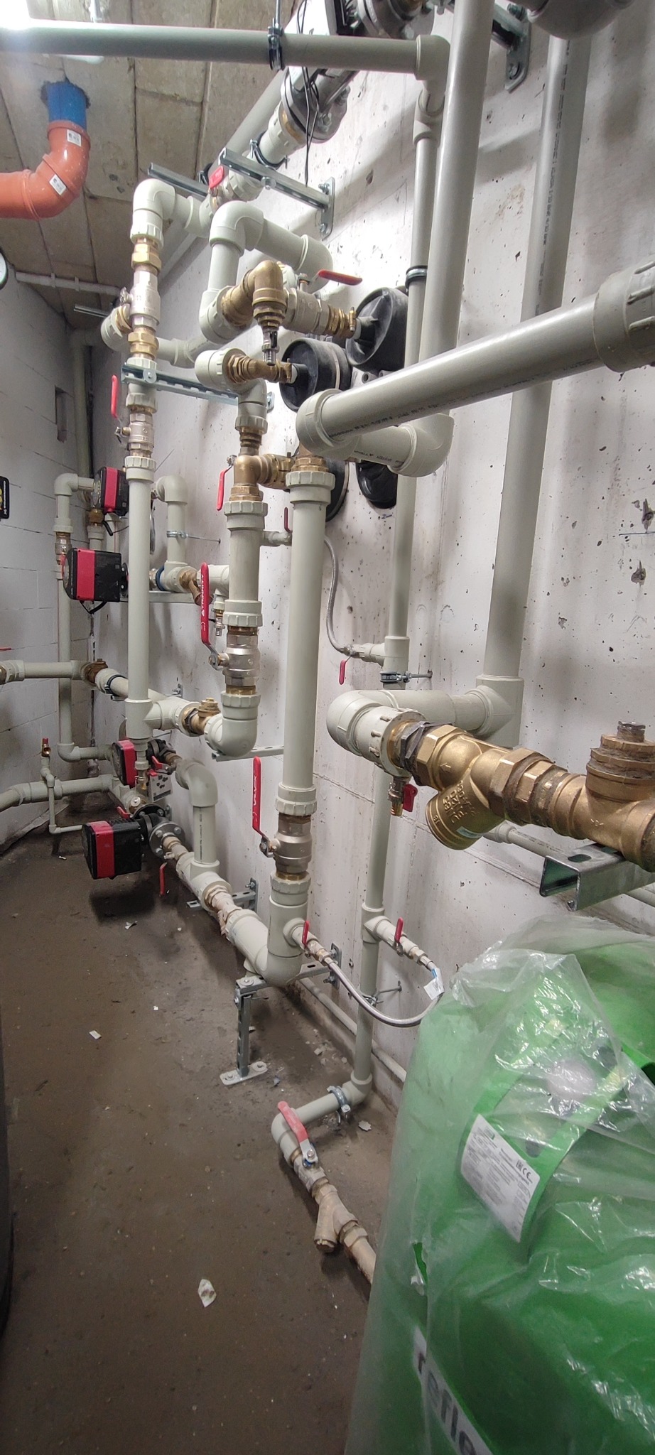 Złożona instalacja hydrauliczna z rur PVC i mosiężnych zaworów, zamontowana na ścianie z widocznymi pompami obiegowymi i zielonym zbiornikiem Reflex w folii.