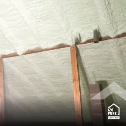 IZOPURE Janusz Walczak - usługi budowlane - Poddasze z drewnianą konstrukcją wypełnione jasną pianką izolacyjną, widoczne rury wentylacyjne i fragment komina. Tekstura pianki przypomina falującą powierzchnię.