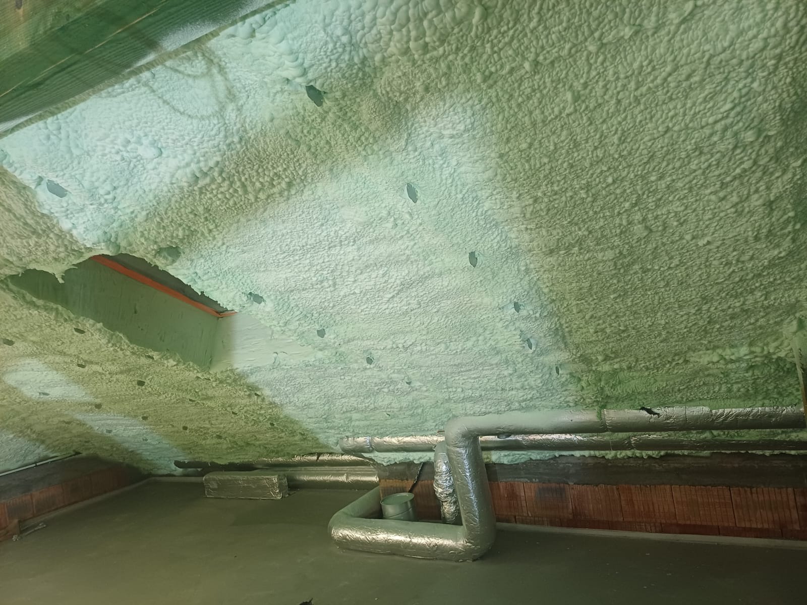 Poddasze po aplikacji pianki PUR, widoczne rury wentylacyjne i fragment ściany z cegły. Aplikacja pianki o jednolitej strukturze i grubości.