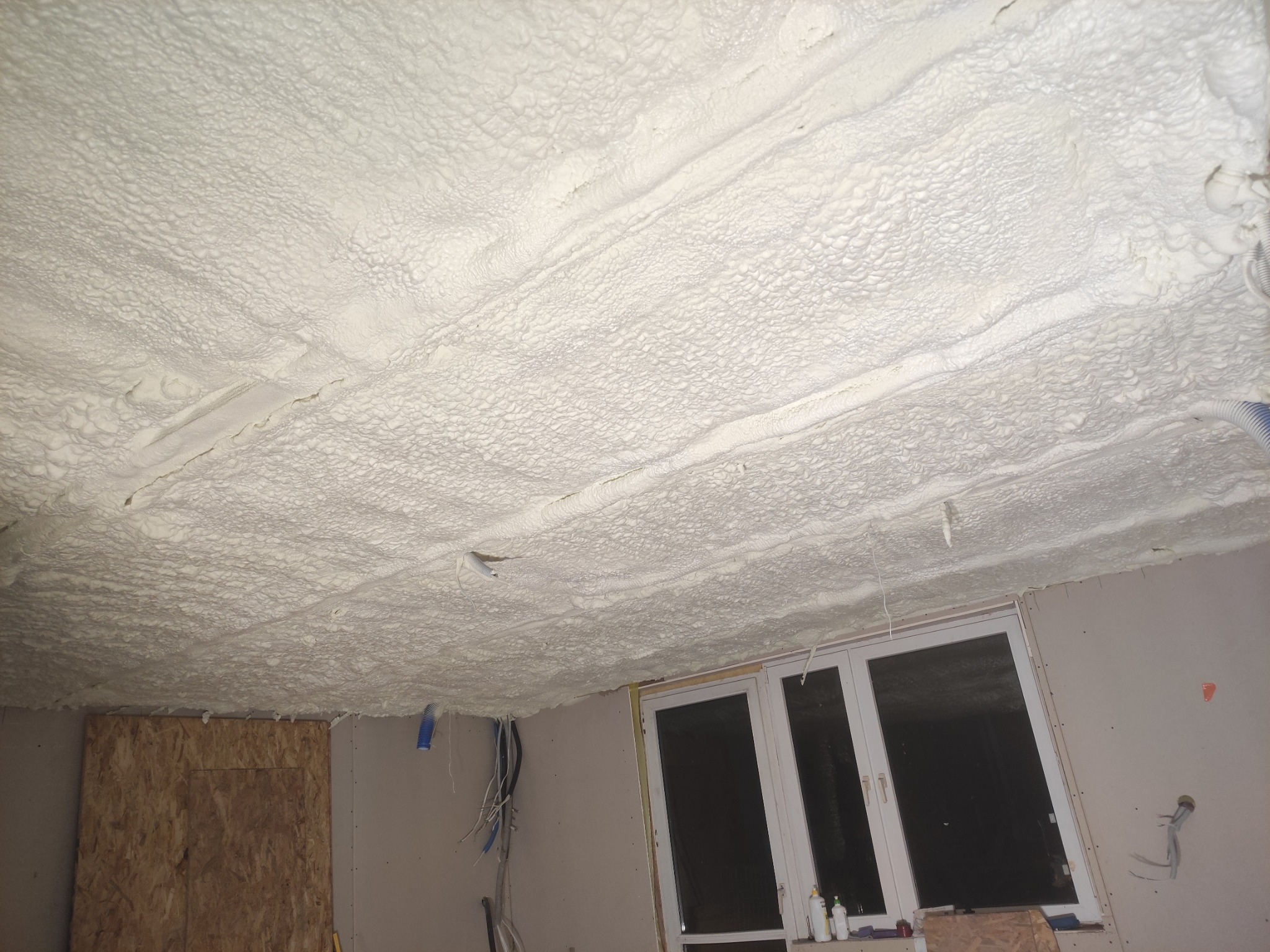 Poddasze po natrysku pianki PUR, widoczne białe, nierównomierne pokrycie izolacyjne. Okno w tle, fragment ściany z instalacją elektryczną.