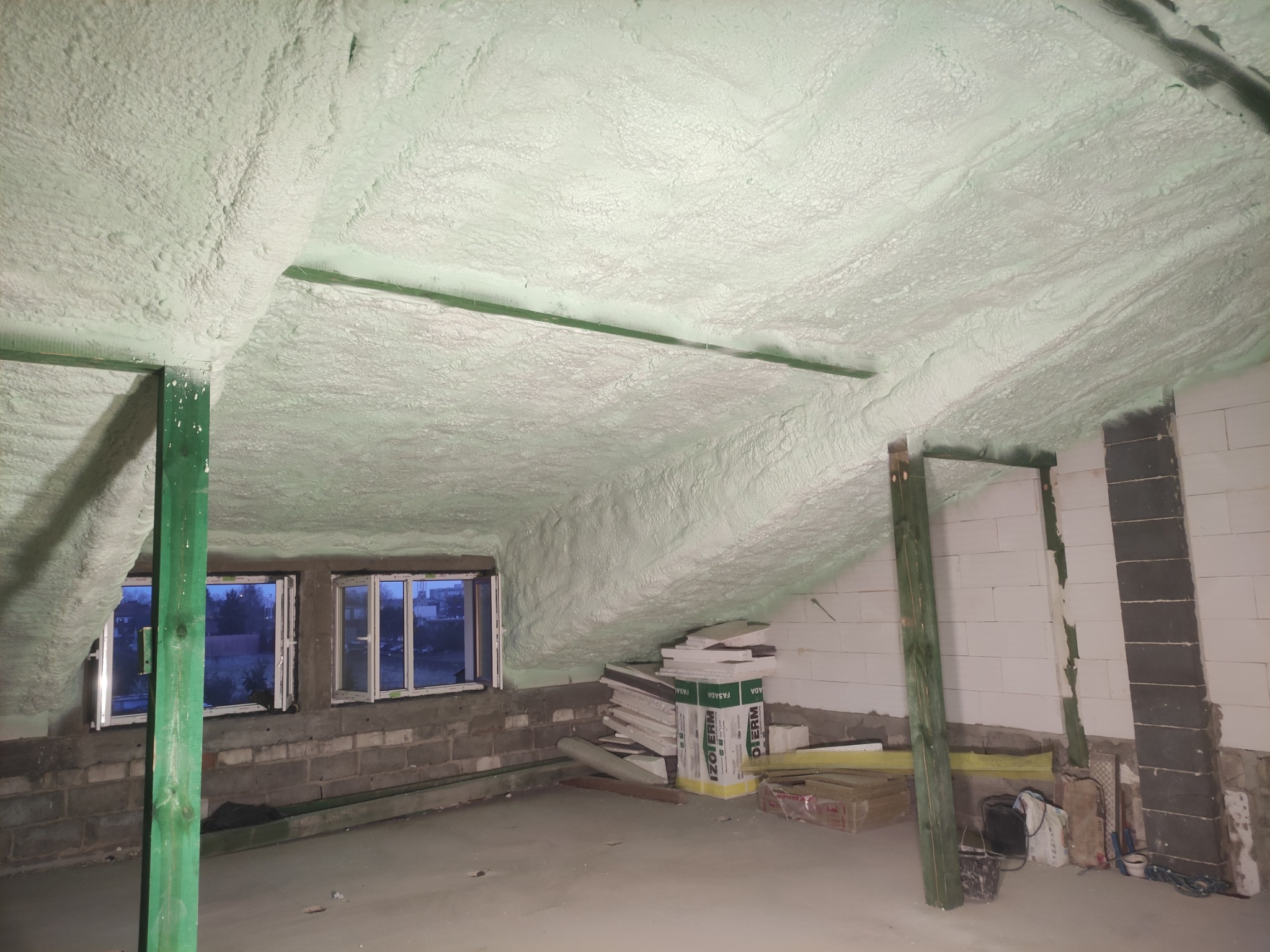 Poddasze z oknami po natrysku pianki PUR, widoczne krokwie i skosy. Na podłodze leżą płyty izolacyjne. Świeża, jasna piana pokrywa całą powierzchnię.