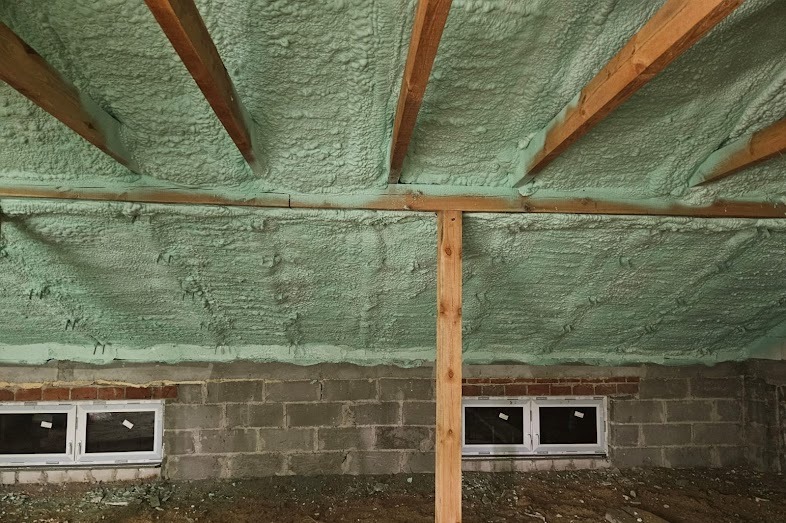 Poddasze z drewnianą konstrukcją i świeżo nałożoną, jasną pianką izolacyjną, widoczne okna w murze z pustaków. Detal wykończenia izolacji.
