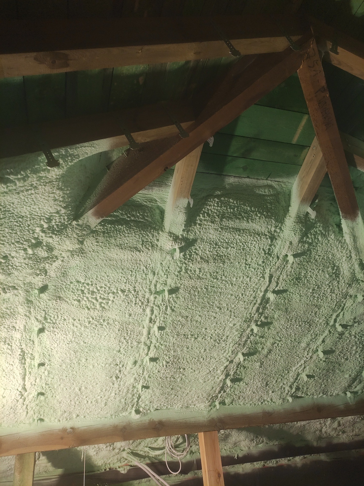 Poddasze z drewnianą konstrukcją pokryte warstwą jasnej pianki izolacyjnej, widoczne elementy mocujące. Ocieplenie wykonane natryskowo.