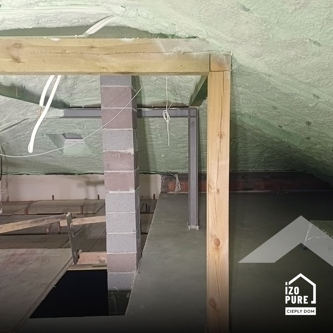 Poddasze po aplikacji pianki PUR, widoczne drewniane elementy konstrukcyjne i fragment ściany z pustaków. Izolacja pokrywa skośny dach i fragment ściany.