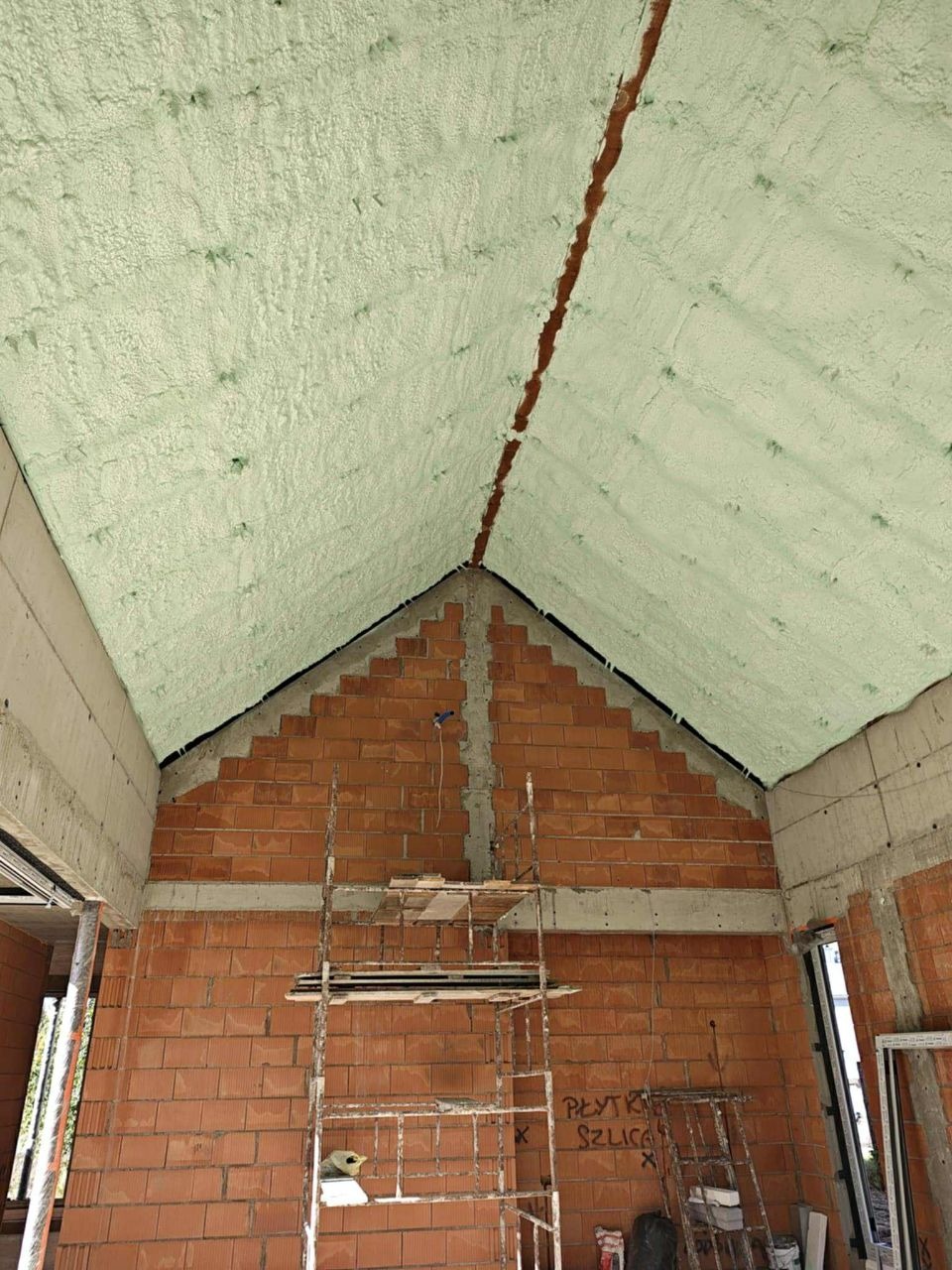 Poddasze z widoczną warstwą izolacji natryskowej, wykonaną pianką. Mur z cegły i rusztowanie w tle. Widoczne detale konstrukcyjne budynku w trakcie prac.