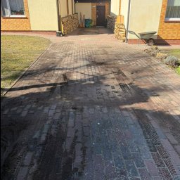 Mycie wysokociśnieniowe Michał Ługowski - Brudna kostka brukowa na podjeździe do domu z numerem 26, widoczne ślady opon i zabrudzenia. Po bokach domu ułożone drewno, w tle błękitne niebo.