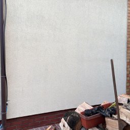 Mycie wysokociśnieniowe Michał Ługowski - Jasna elewacja domu z widocznym fragmentem rynny i stosem drewna opałowego u podstawy ściany. Czerwona ceglana podmurówka i bruk.