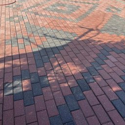 Mycie wysokociśnieniowe Michał Ługowski - Wyczyszczona kostka brukowa w odcieniach czerwieni i szarości, ułożona we wzór. W tle widoczna maszyna czyszcząca i ułożone drewno.