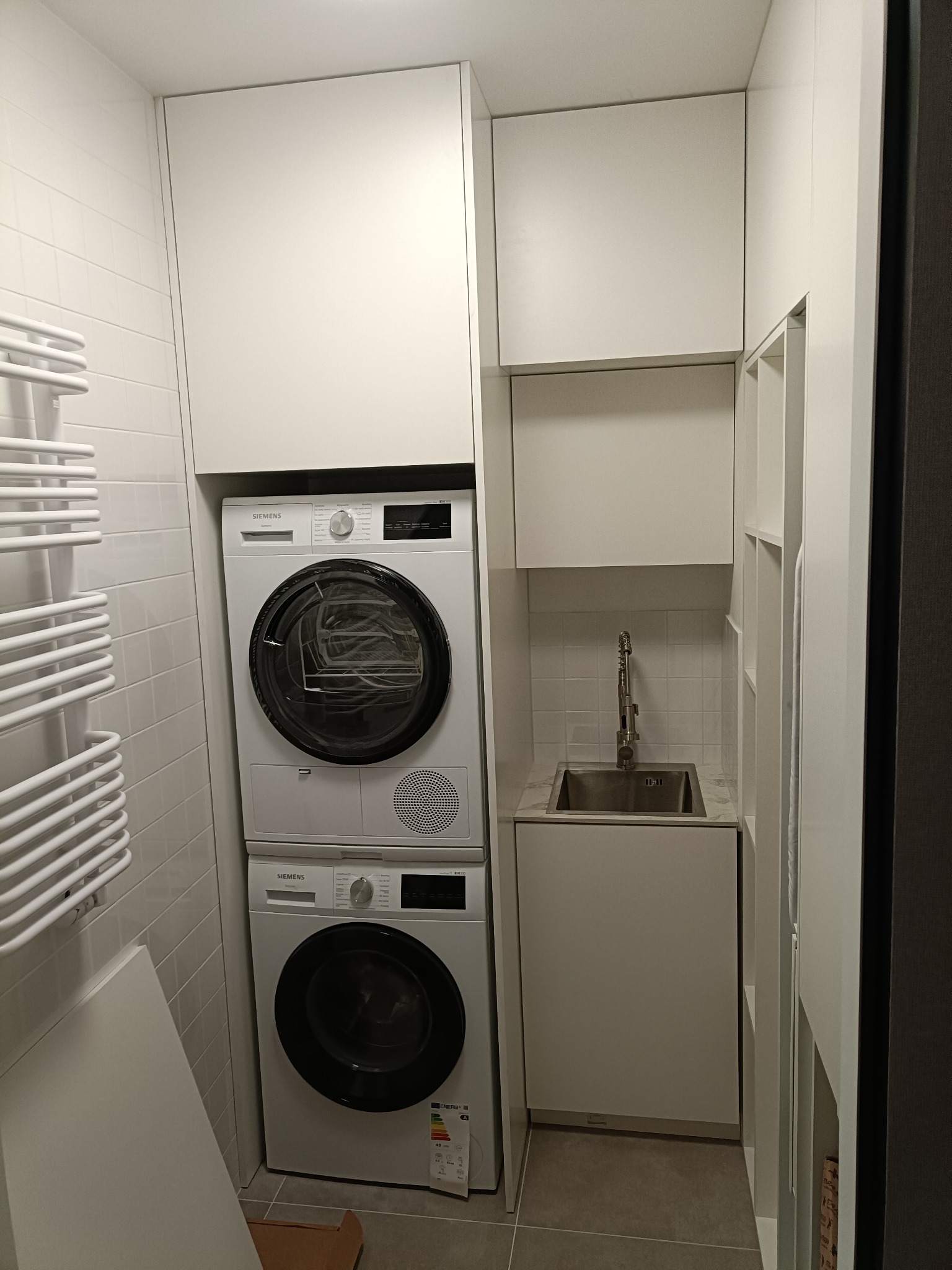 Biała zabudowa w łazience: pralka i suszarka Siemens w kolumnie, szafki wiszące, umywalka ze stali nierdzewnej, grzejnik drabinkowy na tle białych płytek.