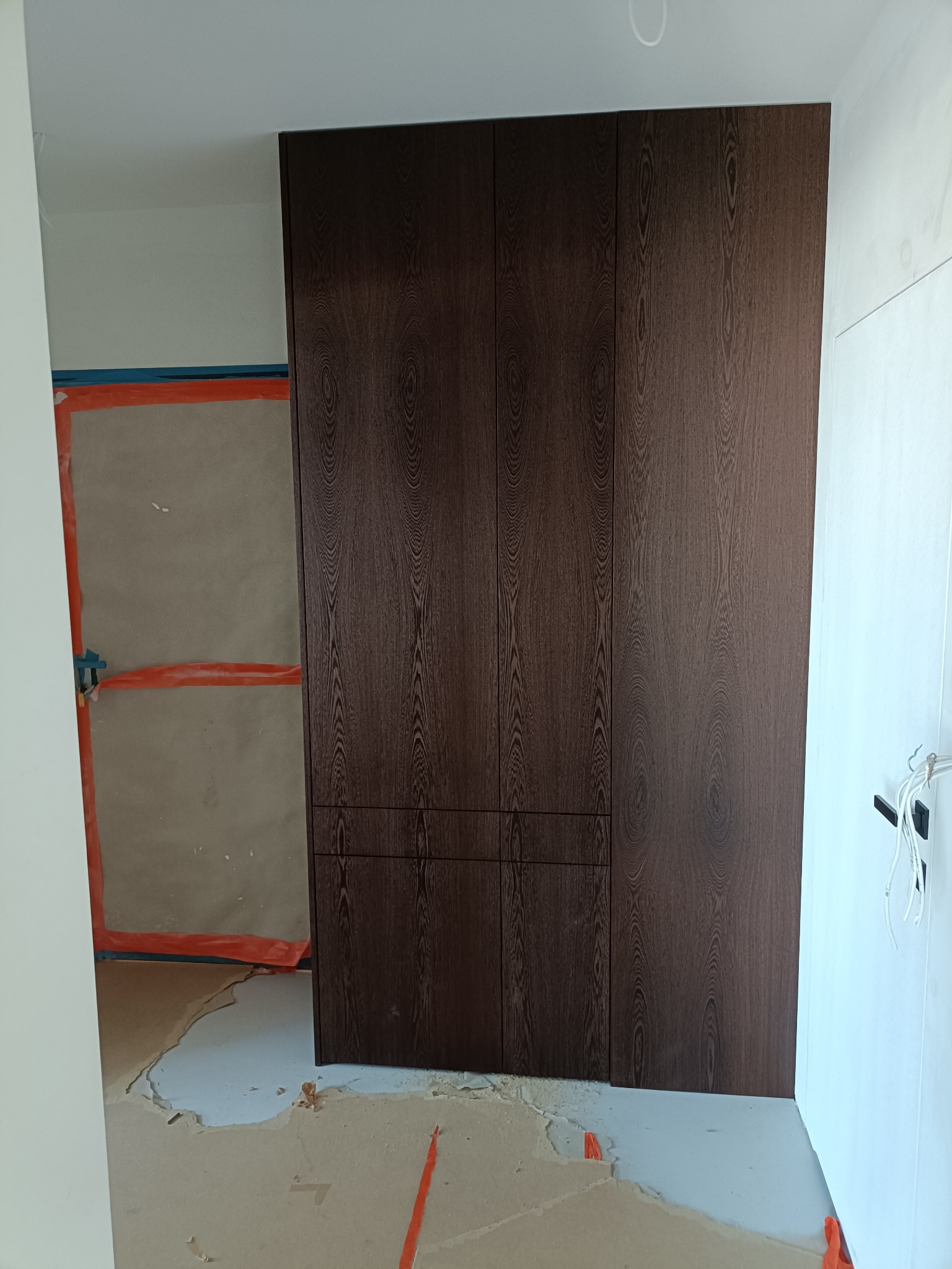 Ciemnobrązowa szafa wnękowa z wyraźnym rysunkiem drewna, w surowym wnętrzu w trakcie remontu. Widoczne zabezpieczenia ścian i podłogi.