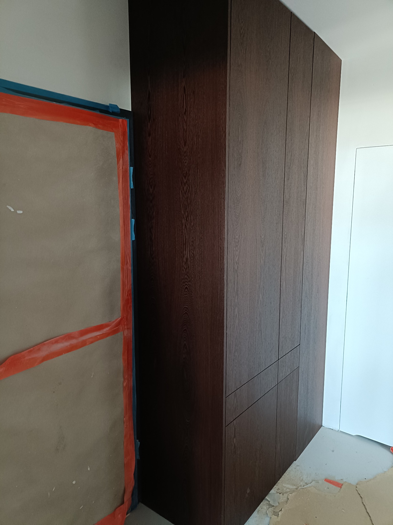 Ciemnobrązowa szafa wnękowa z widoczną strukturą drewna, zamontowana w pomieszczeniu w trakcie wykańczania, z zabezpieczonymi folią drzwiami obok.