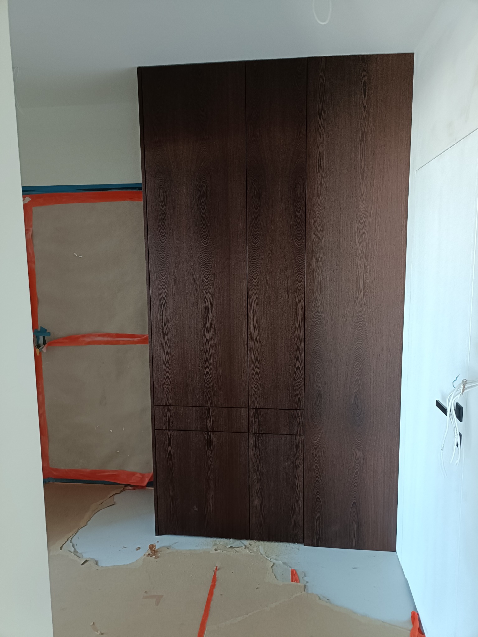 Ciemnobrązowa szafa wnękowa z widoczną strukturą drewna, zamontowana w pomieszczeniu w trakcie remontu. Na podłodze i ścianach widoczne ślady prac wykończeniowych.