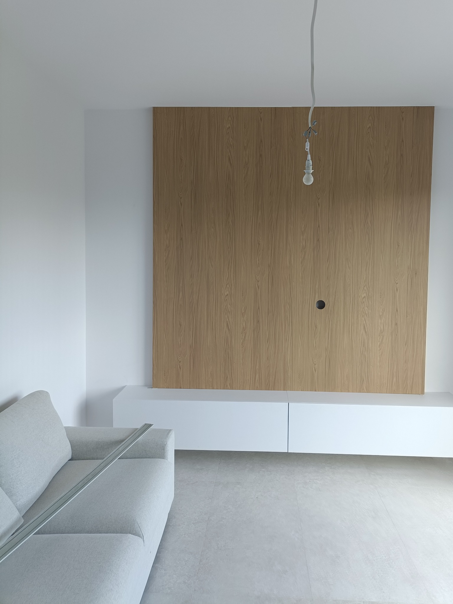 Minimalistyczny salon: drewniana ściana TV z otworem na kable, biała szafka RTV, szara sofa i wisząca żarówka na tle jasnych ścian i podłogi.