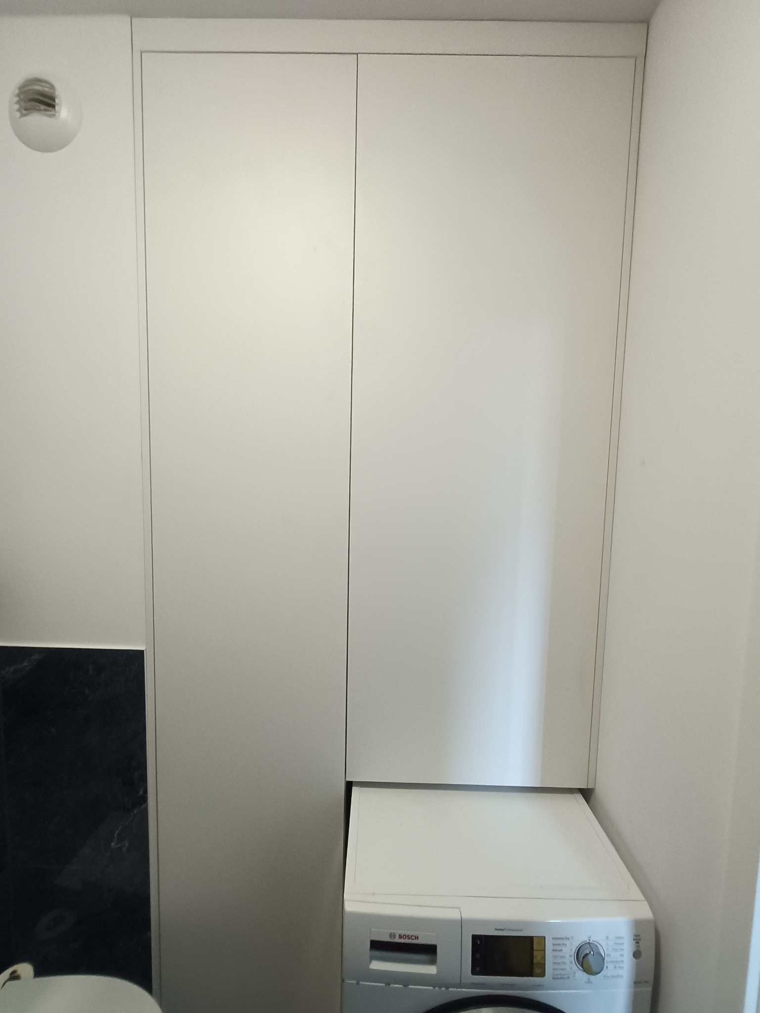 Biała szafa wnękowa z pralką Bosch w łazience. Minimalistyczny design, gładkie fronty, oświetlenie sufitowe. Funkcjonalne rozwiązanie do małych przestrzeni.