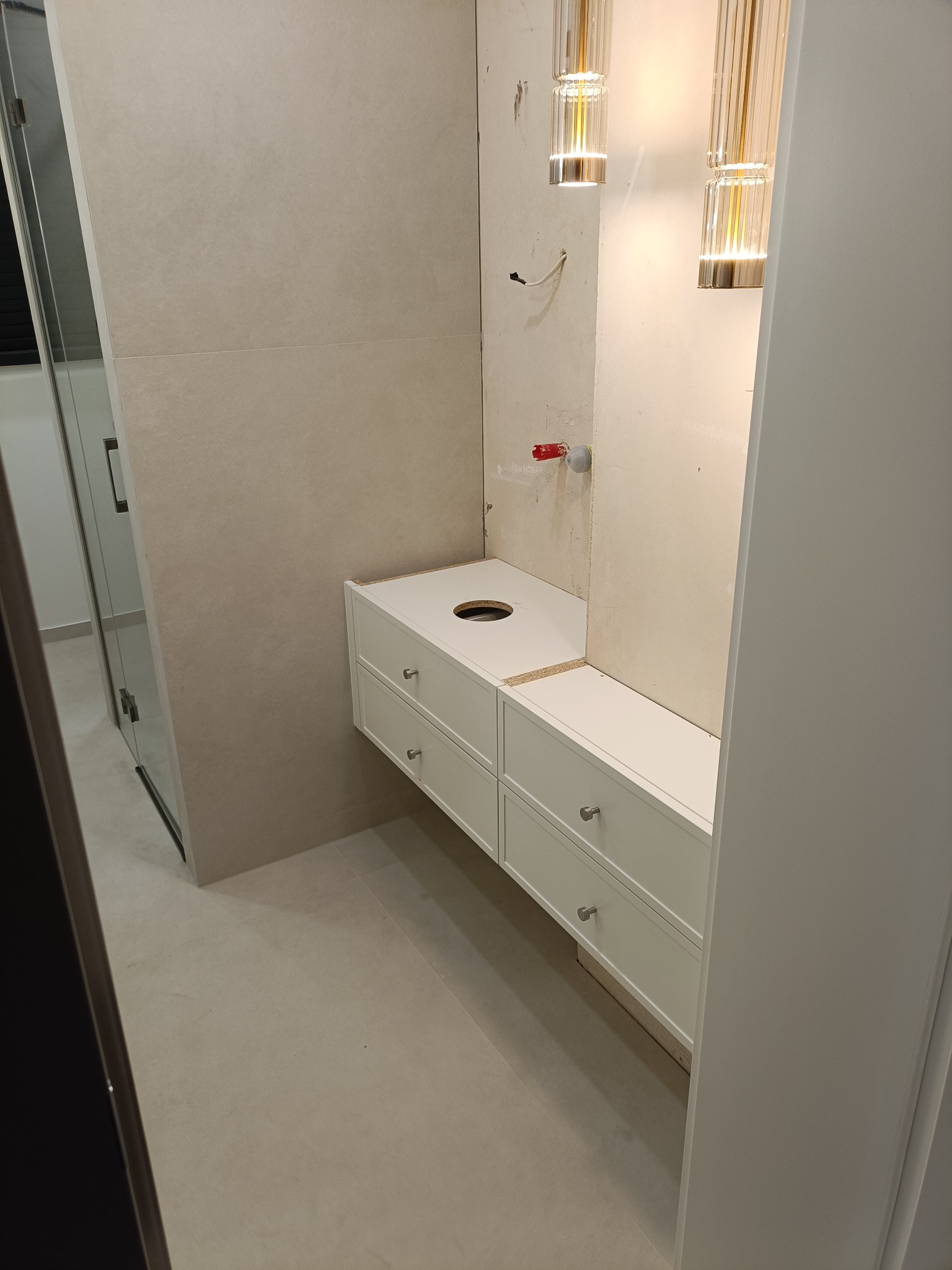 Biała szafka łazienkowa z czterema szufladami, bez blatu i umywalki, zamontowana na ścianie z beżowymi płytkami. Widoczne lampy wiszące i fragment kabiny prysznicowej.