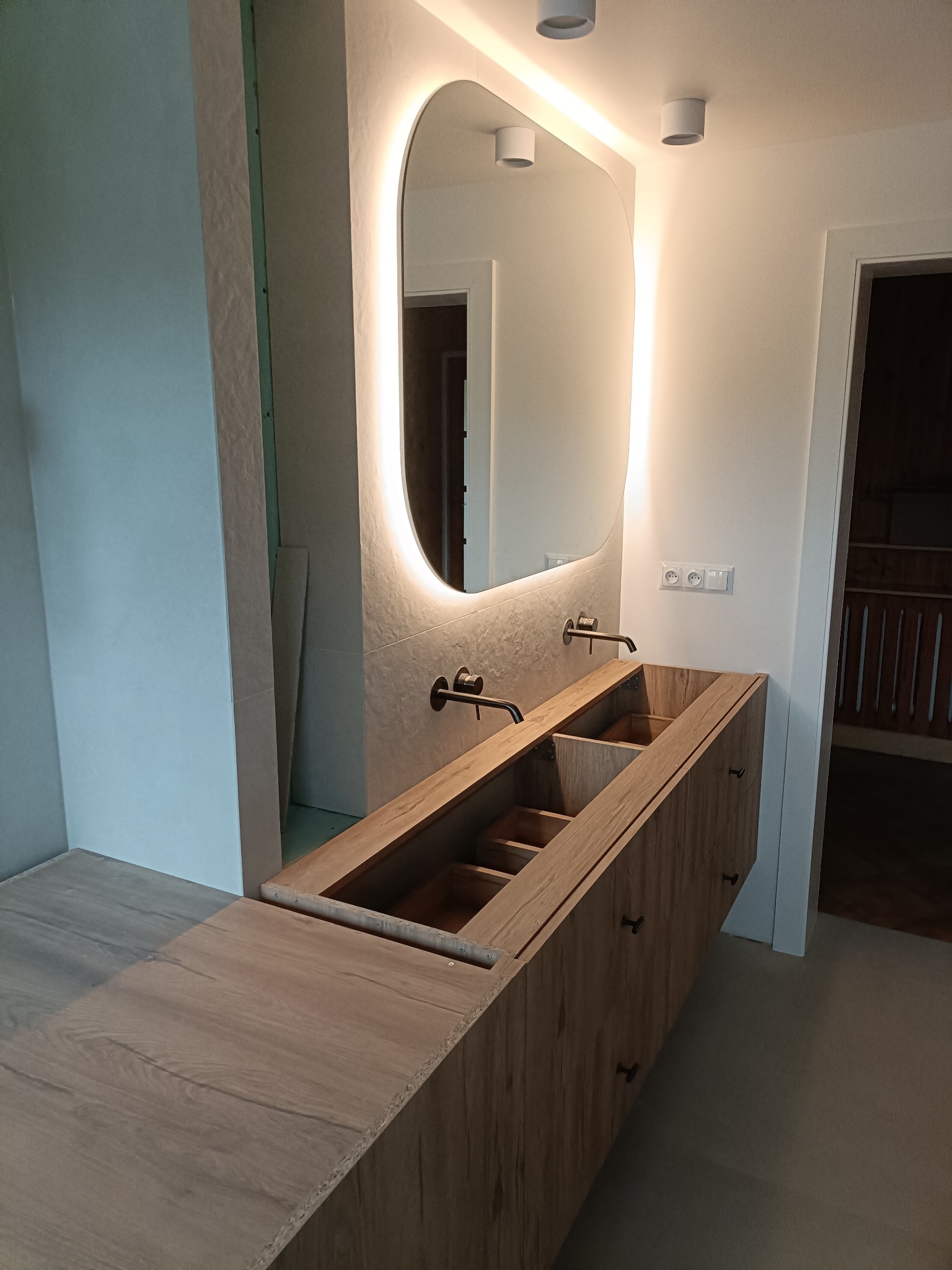 Nowoczesna łazienka w trakcie montażu: drewniana szafka pod umywalkę z czarną armaturą i podświetlanym lustrem owalnym. Widoczne elementy instalacji i surowa ściana.