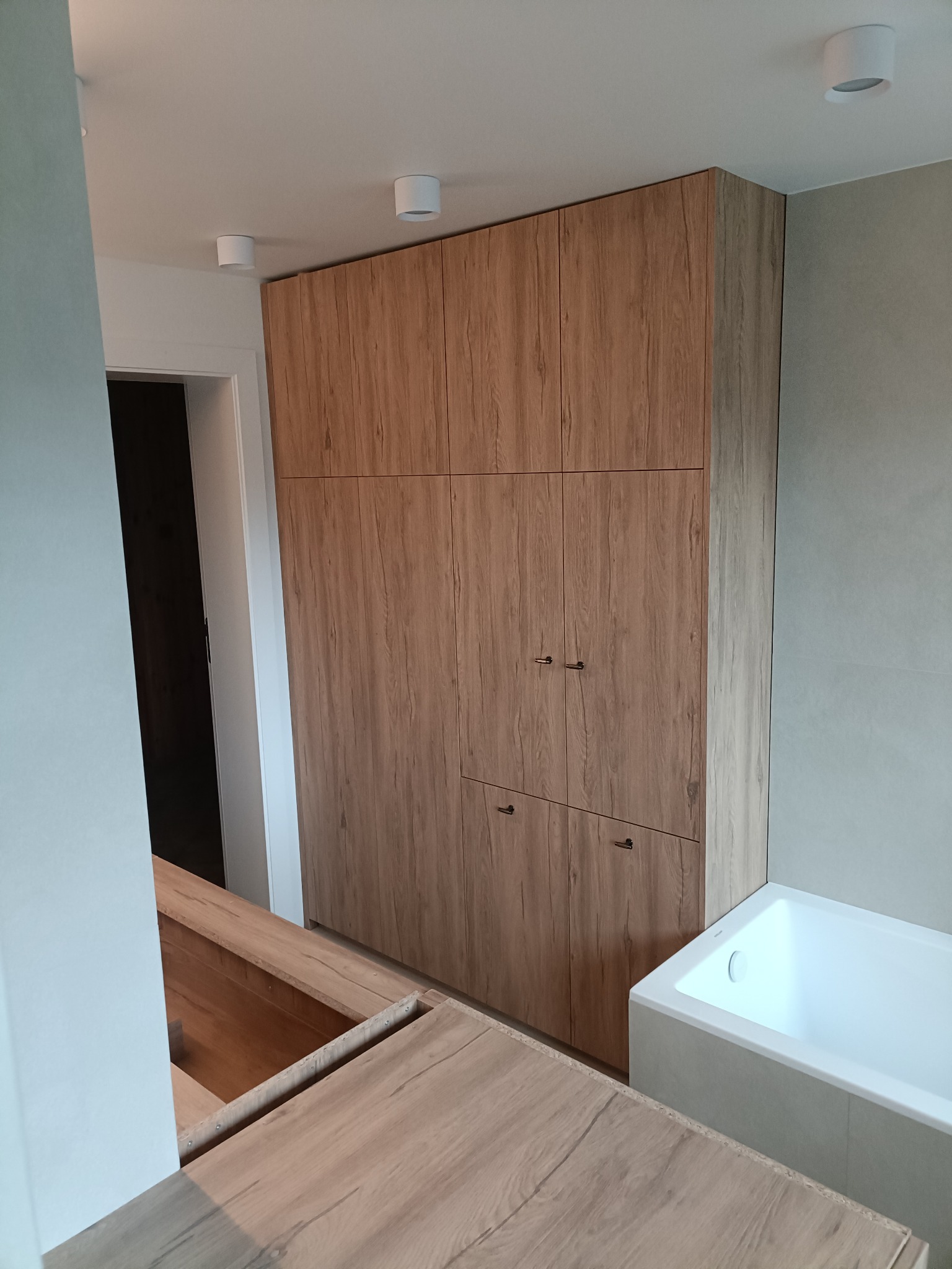 Wysoka zabudowa z drewnopodobnych frontów w łazience, zintegrowana z wanną. Minimalistyczne uchwyty, punktowe oświetlenie sufitowe. Częściowo widoczny blat.