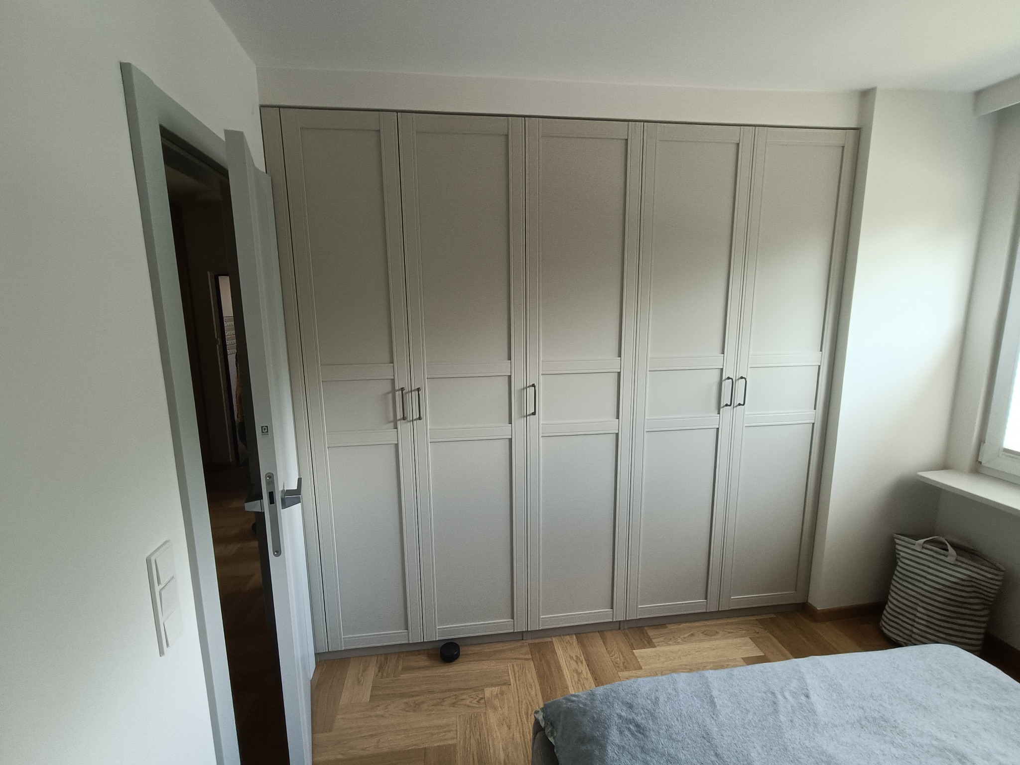 Jasnoszara szafa wnękowa z sześcioma drzwiami, zamontowana w sypialni w Warszawie. Parkiet w jodełkę, widoczny fragment łóżka i okno z koszem na pranie.