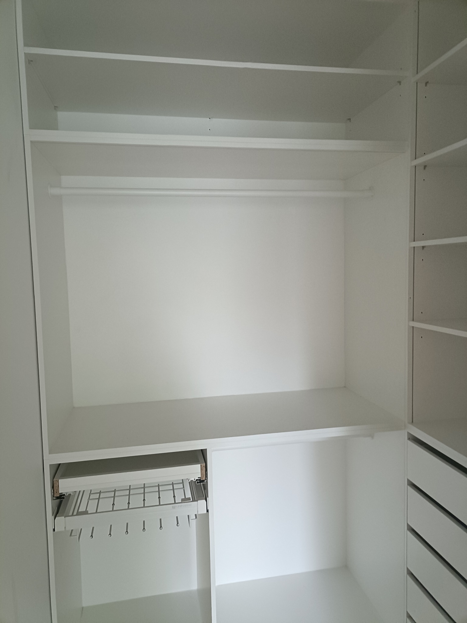 Wnętrze białej, nowoczesnej szafy wnękowej z drążkiem na ubrania i wysuwanym wieszakiem na spodnie. Widoczne półki i szuflady po prawej stronie.