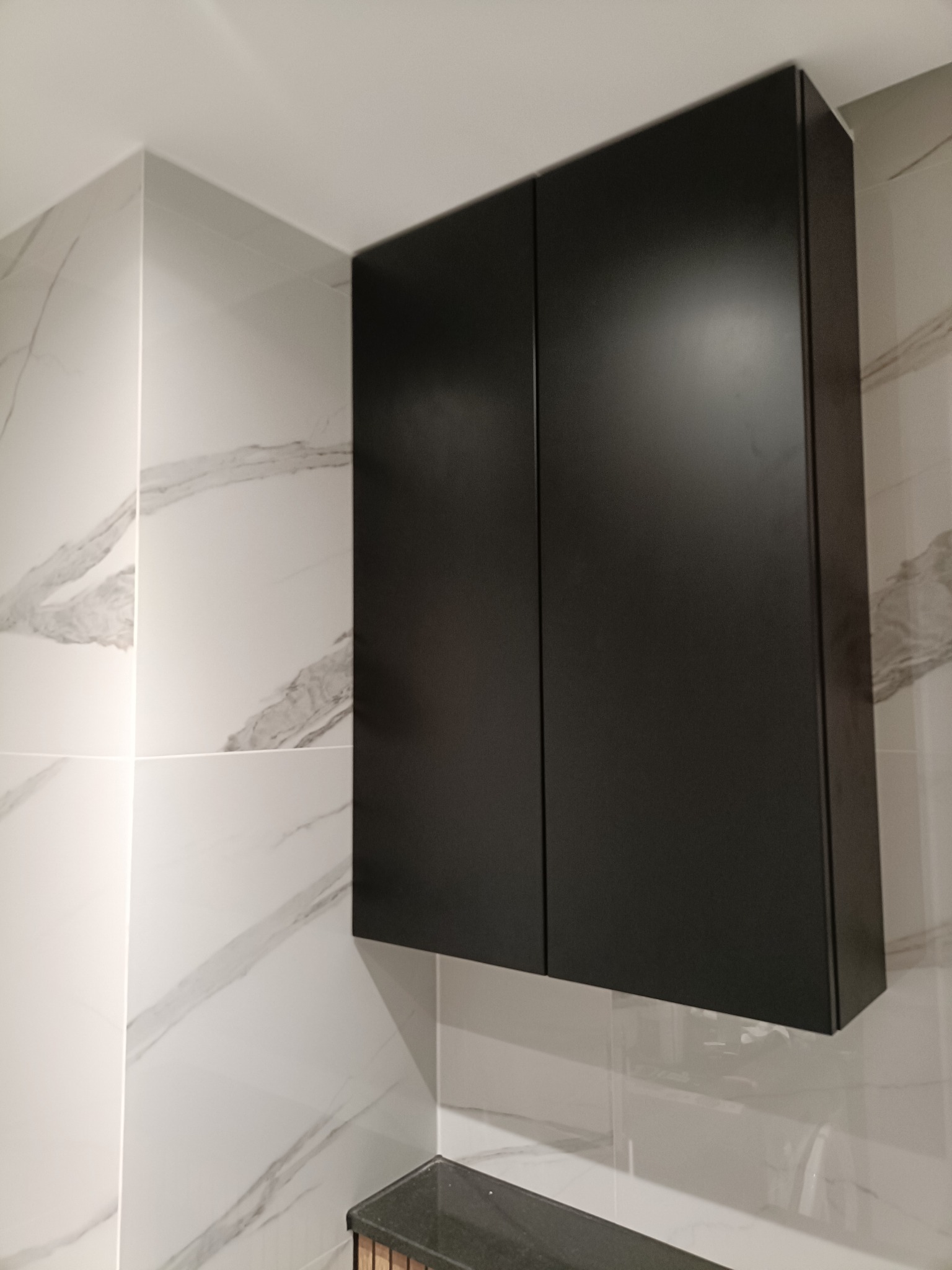 Czarna szafka wisząca z dwoma drzwiczkami, zamontowana na ścianie z marmurowymi płytkami w łazience. Minimalistyczny design, nowoczesne wykończenie.