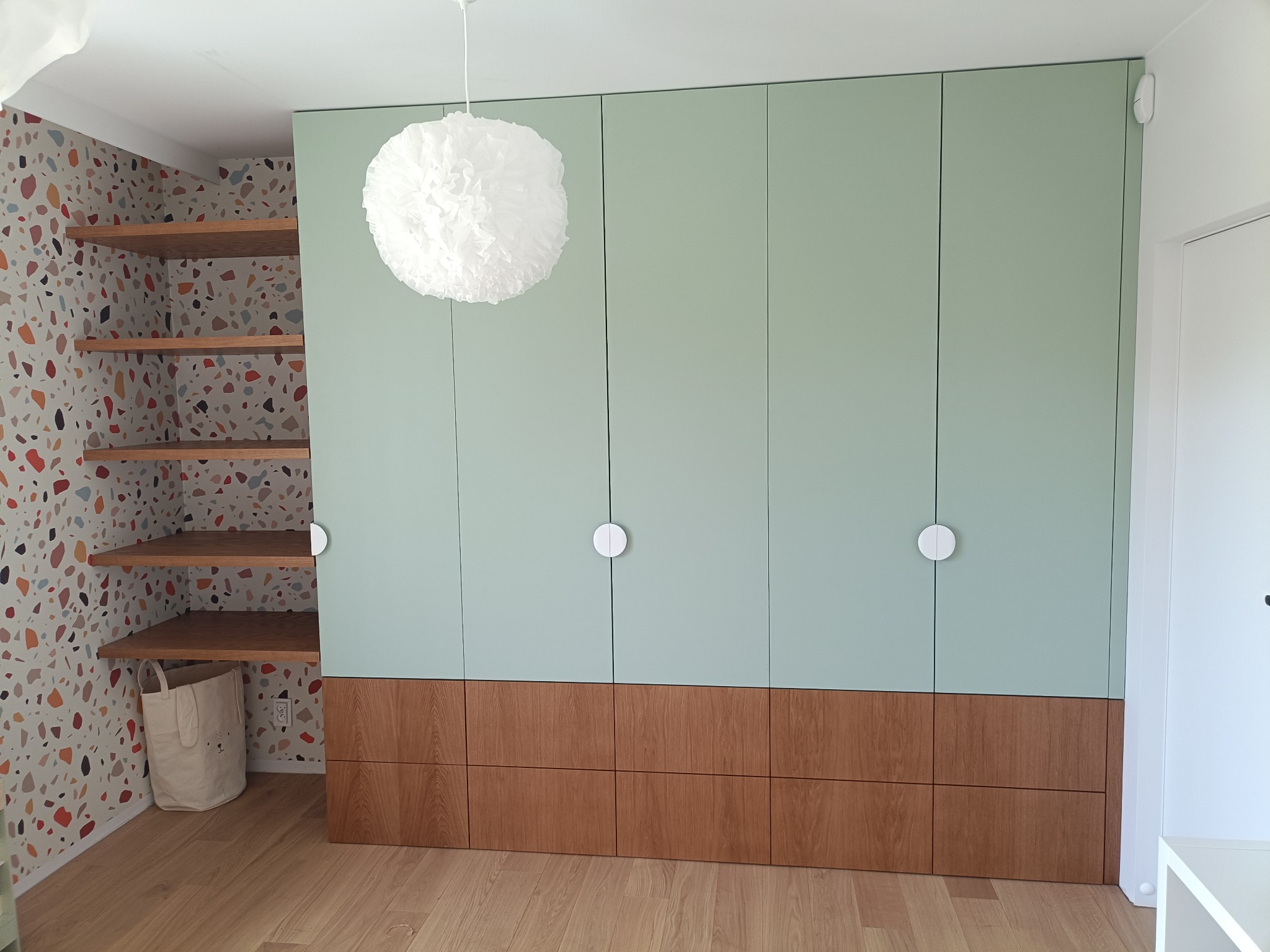 Pistacjowa szafa wnękowa z drewnianą podstawą i okrągłymi uchwytami, uzupełniona otwartymi półkami z drewnianymi blatami i wzorzystą tapetą w tle.