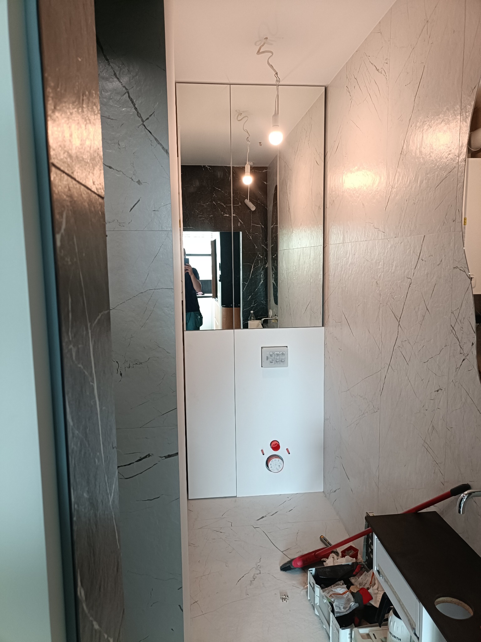 Nowoczesna łazienka w trakcie remontu: marmurowe płytki, biała zabudowa, lustro z odbiciem osoby. Widoczne narzędzia i akcesoria remontowe. Oświetlenie sufitowe.