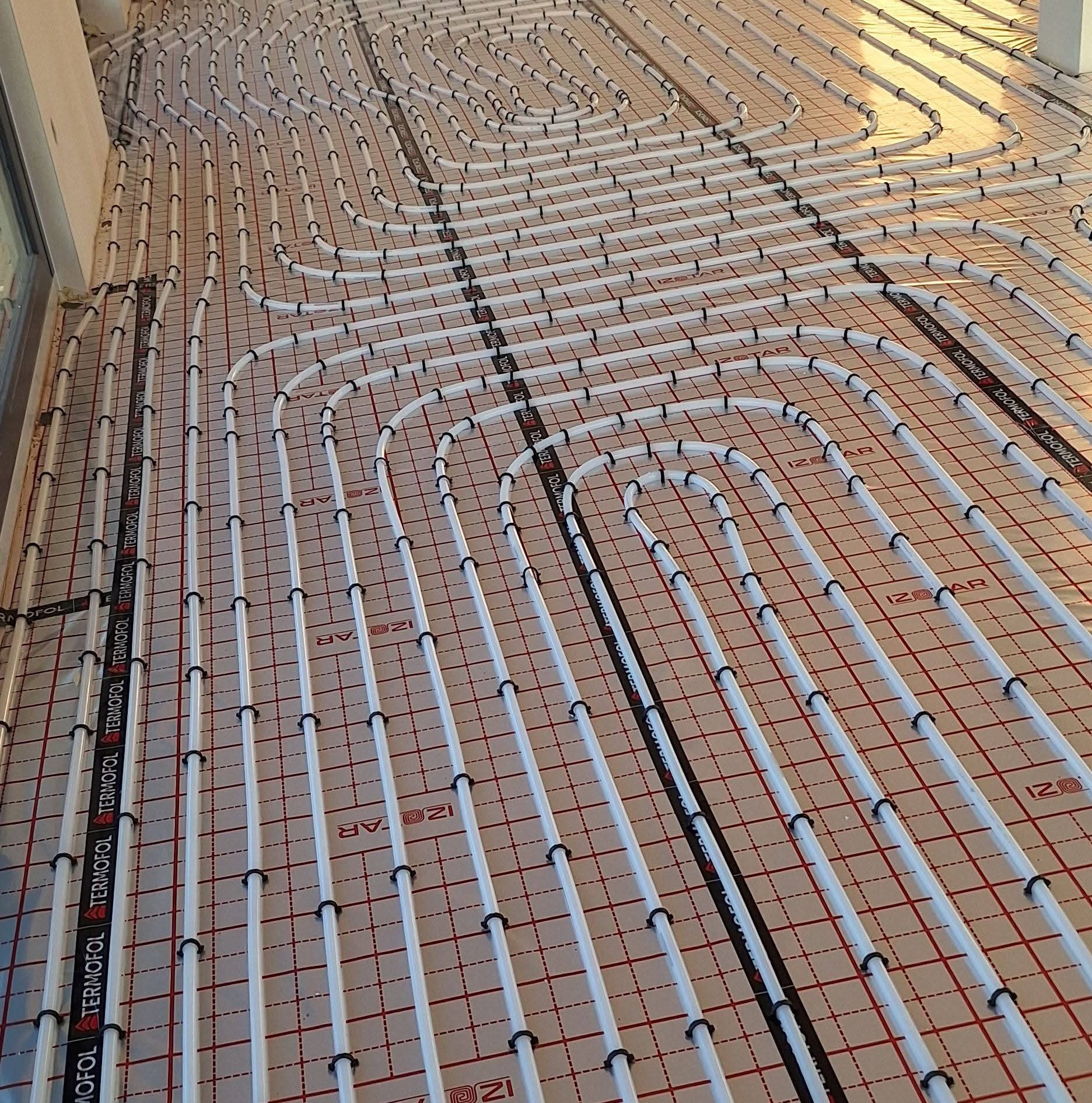 Białe rury ogrzewania podłogowego ułożone na czerwonej siatce, przymocowane czarnymi uchwytami. Widoczne napisy 'TERMOFOL' i 'IZOTAR' na elementach instalacji.