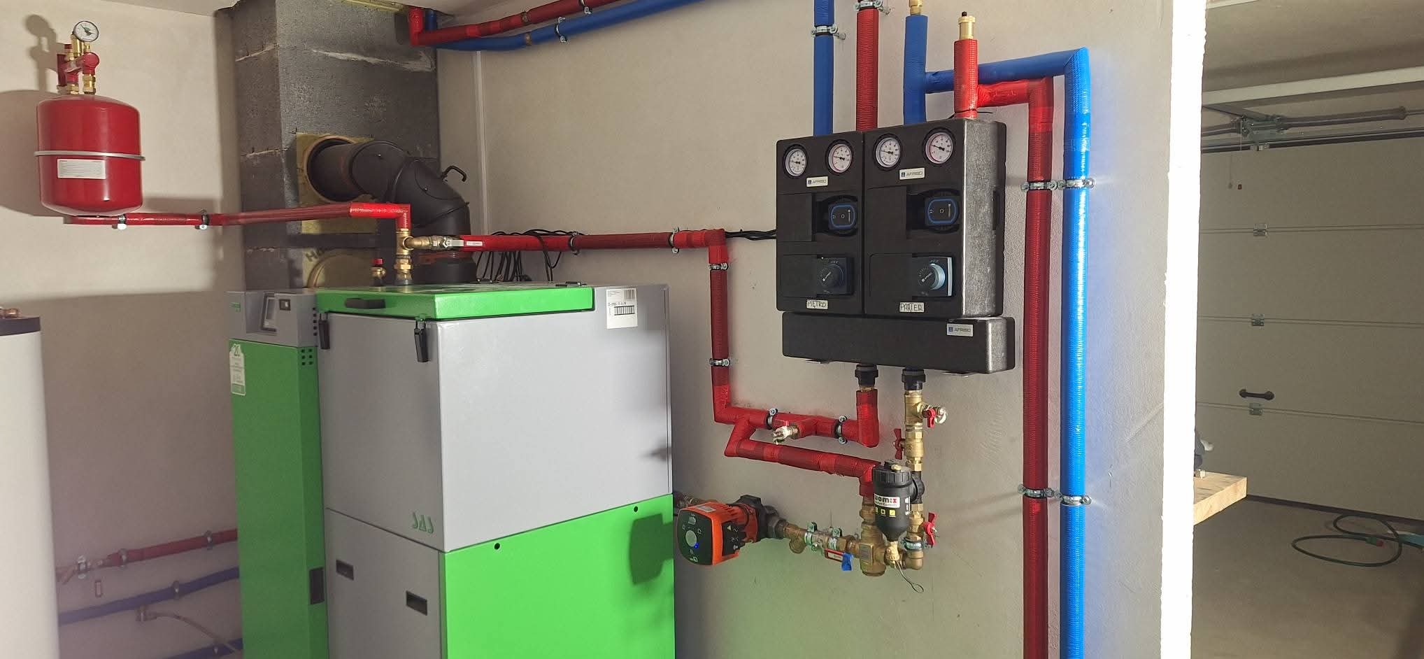 Nowoczesna kotłownia z piecem na pellet, zbiornikiem wyrównawczym i rozdzielaczem z manometrami. Instalacja z rur w kolorach czerwonym i niebieskim.