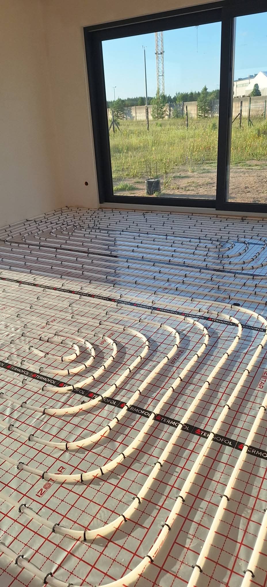 Białe rury ogrzewania podłogowego ułożone na folii z czerwoną siatką w jasnym pomieszczeniu z dużym oknem, przez które widać zieloną trawę i budynki w oddali.