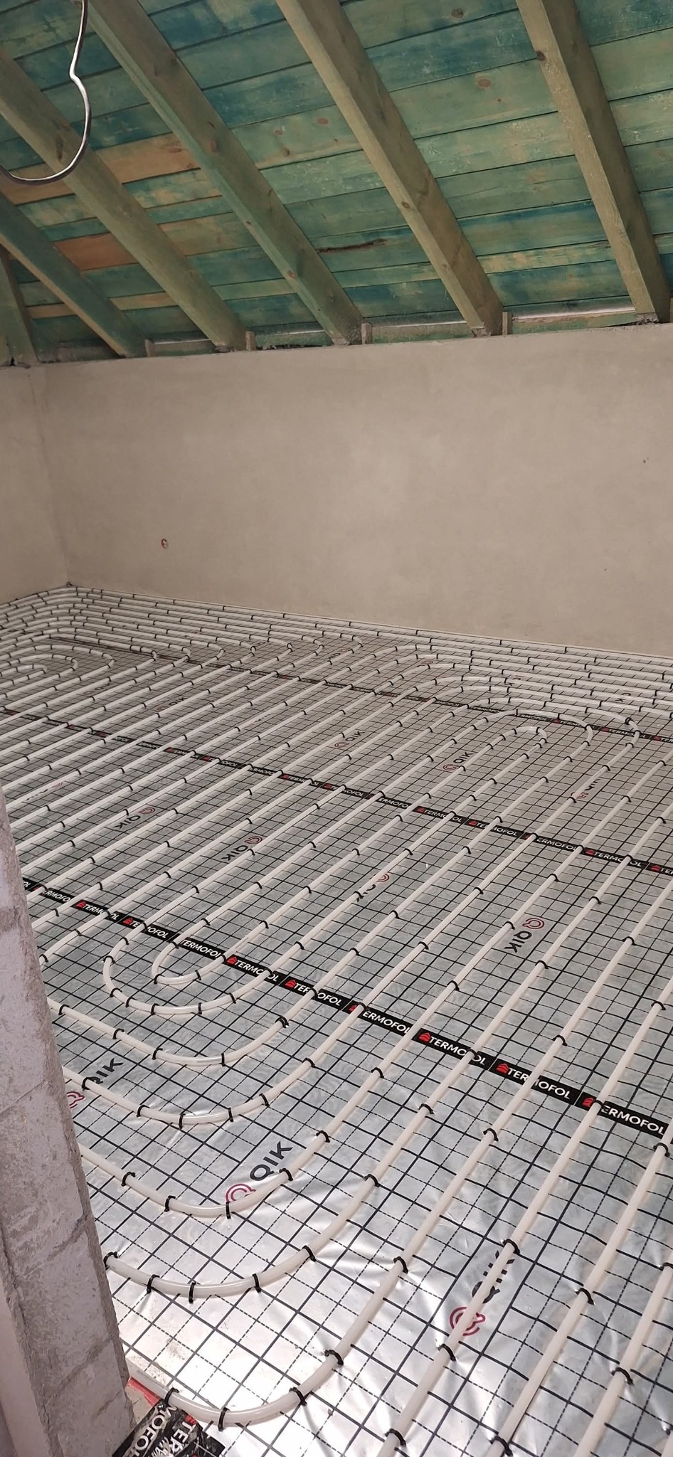 Białe rury ogrzewania podłogowego ułożone na srebrnej folii z czarną siatką, widok z góry, surowy stan wykończenia wnętrza z drewnianym stropem.