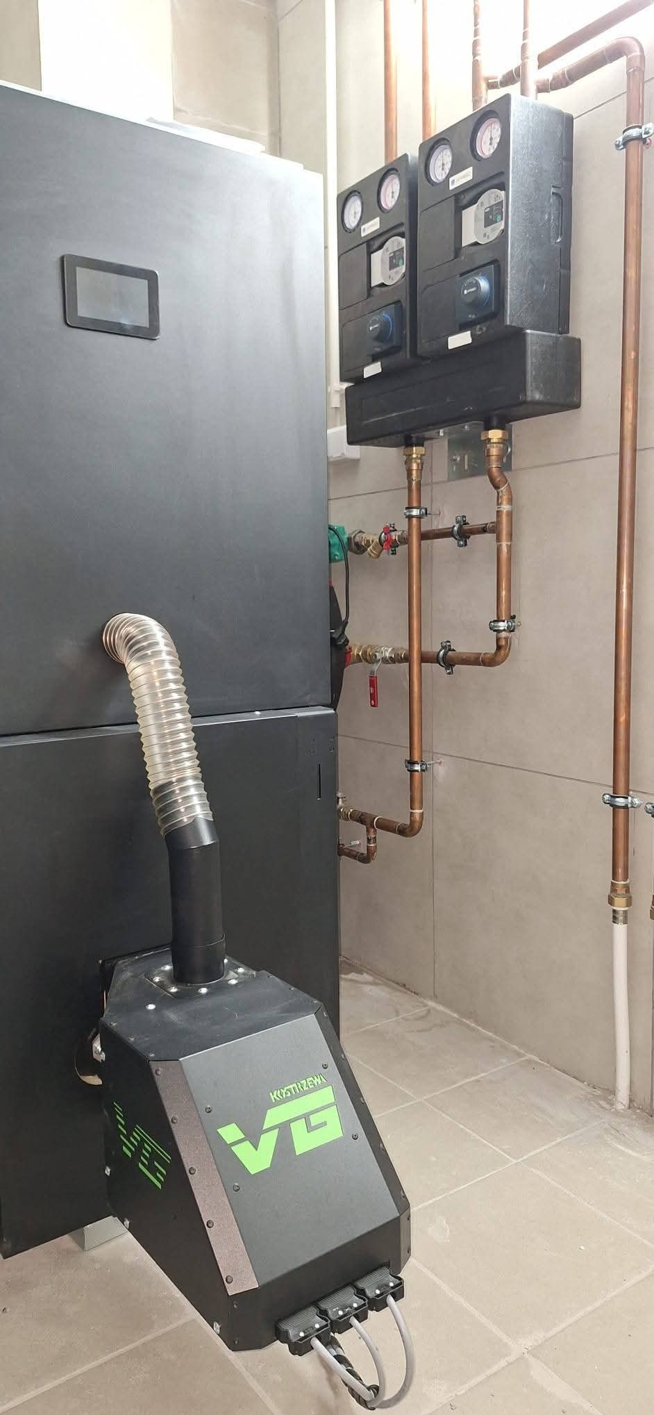 Nowoczesny piec CO marki Kostrzewa VG z podajnikiem, zainstalowany w kotłowni z widoczną instalacją hydrauliczną i osprzętem kontrolnym na ścianie.