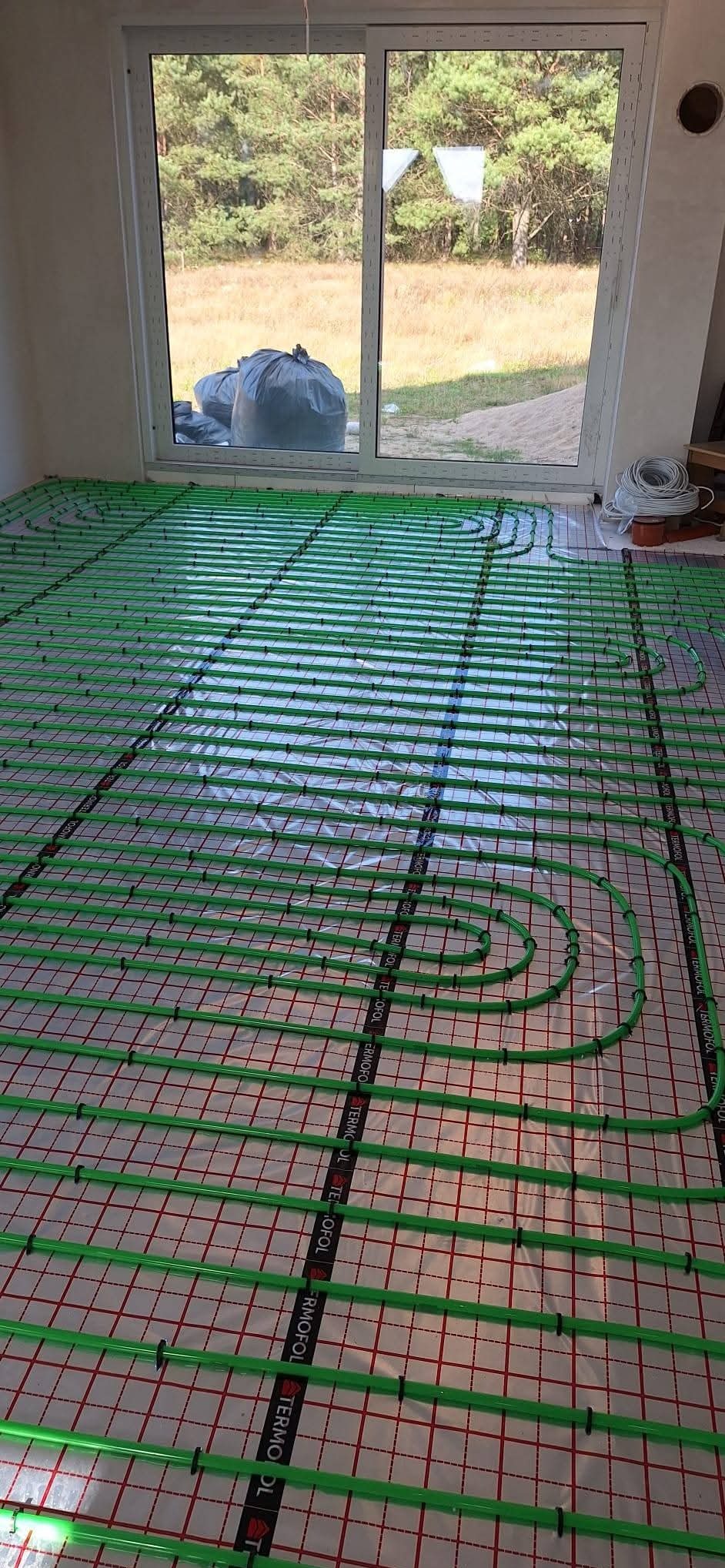 Zielone rury ogrzewania podłogowego ułożone na folii z czerwoną siatką, widok z góry, w tle okno z widokiem na zewnątrz. Instalacja w trakcie realizacji.