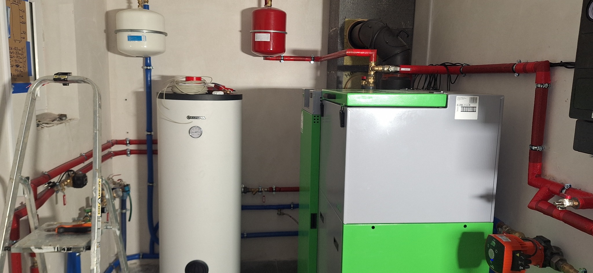 Kotłownia z piecem CO na pellet, zbiornikiem buforowym Kospel i instalacją z rur miedzianych. Widoczna drabina i schemat instalacji na ścianie.