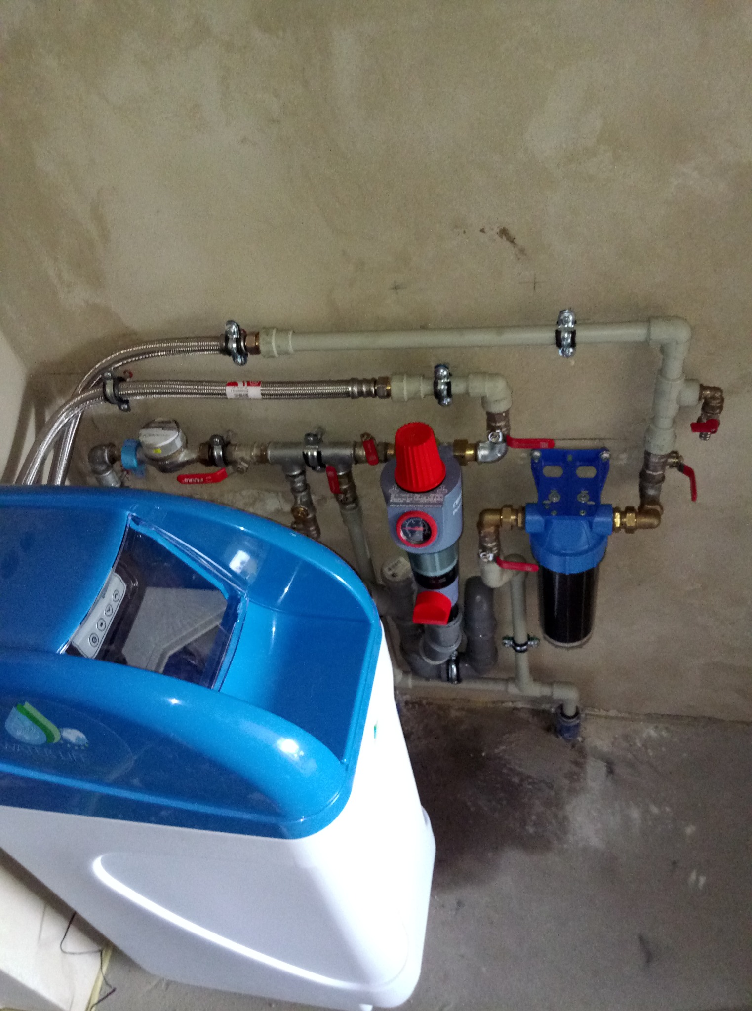 Biało-niebieski zmiękczacz wody podłączony do instalacji hydraulicznej z filtrem i reduktorem ciśnienia. Widoczne rury PCV i metalowe złączki.