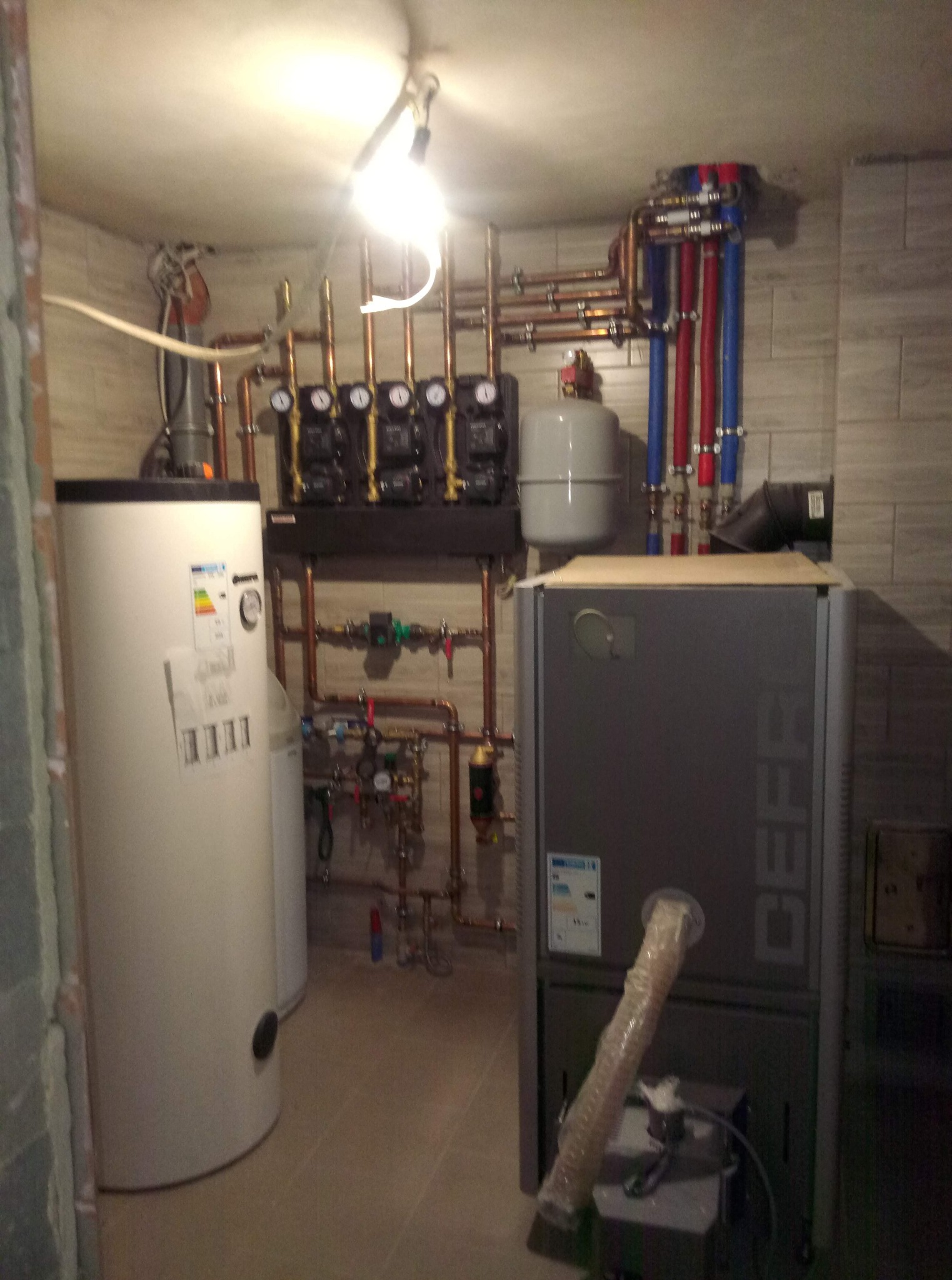 Złożona instalacja grzewcza w kotłowni: piec, zbiornik, pompy obiegowe i rury miedziane z oznaczeniami kolorystycznymi. Widoczne elementy montażowe i oświetlenie robocze.