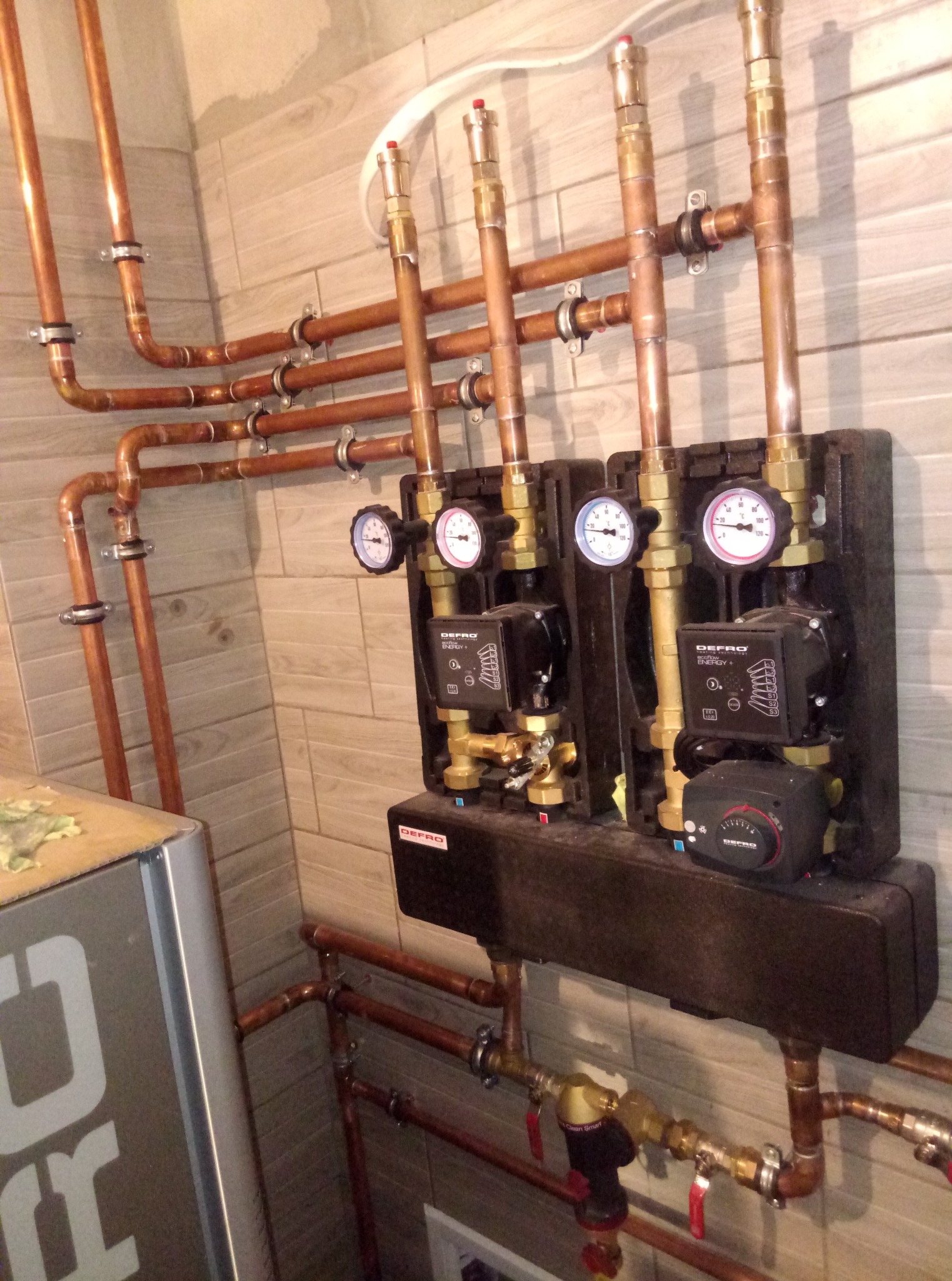 Kompleksowa instalacja grzewcza z miedzianymi rurami, manometrami i pompami obiegowymi Defro na tle płytek imitujących drewno. Precyzyjne wykonanie.