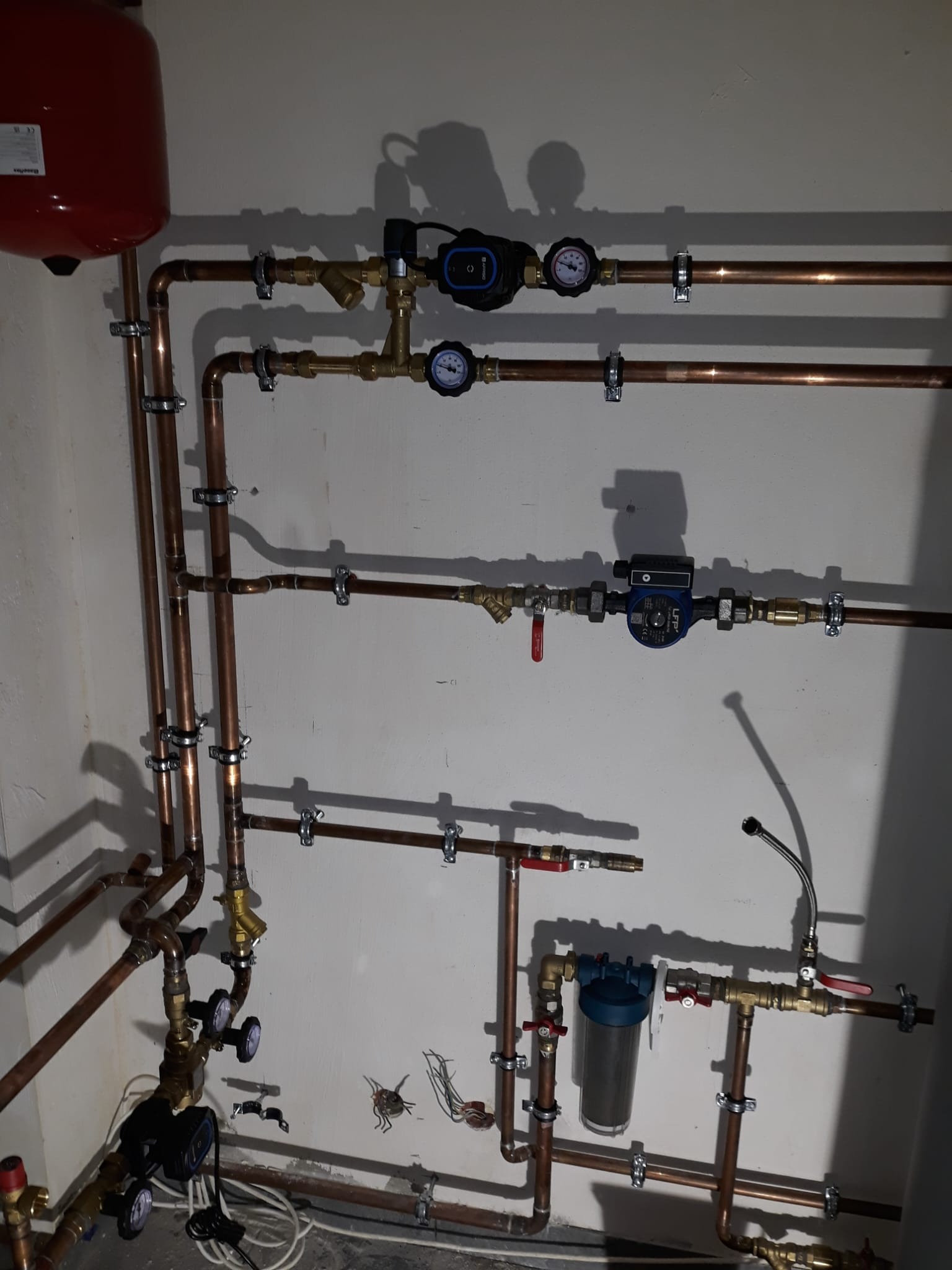 Kompleksowa instalacja hydrauliczna z miedzianych rur, zaworów, manometrów i pompy obiegowej zamontowana na ścianie. Widoczne liczne mocowania i elementy instalacji.