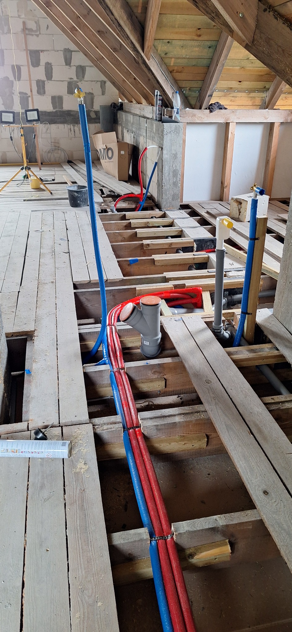 Instalacja wodna i kanalizacyjna w trakcie montażu na poddaszu. Widoczne rury w kolorze niebieskim i czerwonym, szare rury kanalizacyjne, drewniana konstrukcja podłogi.