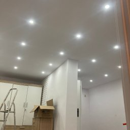 ART-G - Jasne wnętrze po malowaniu z równomiernie rozmieszczonymi punktowymi lampami sufitowymi. Widoczny fragment drabiny i mebli w trakcie prac wykończeniowych.