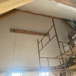 ART-G - Skośny sufit poddasza z płyt kartonowo-gipsowych, widoczne drewniane belki konstrukcyjne i rusztowanie. Praca w toku, widok z dołu.
