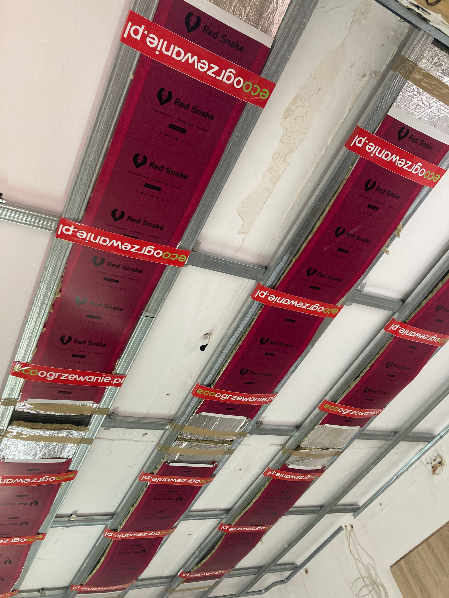Montaż folii grzewczej Red Snake na metalowym stelażu sufitowym, widoczne izolacje i elementy instalacji. Prace wykończeniowe w trakcie realizacji.