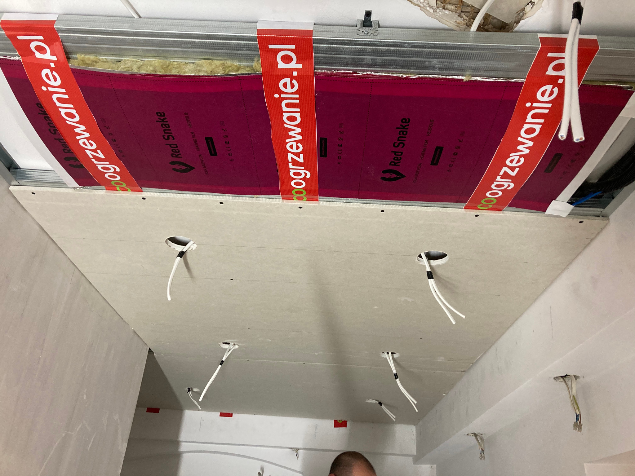 Montaż sufitu podwieszanego z płyt karton-gips z widoczną instalacją grzewczą Red Snake i wyprowadzonymi kablami elektrycznymi. Etap prac wykończeniowych.