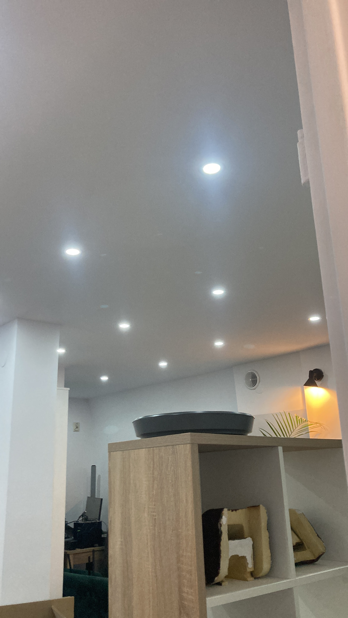 Jasne wnętrze z rzędem punktowych lamp sufitowych. Widoczny regał z dekoracjami i fragment pomieszczenia biurowego w tle. Minimalistyczny design.