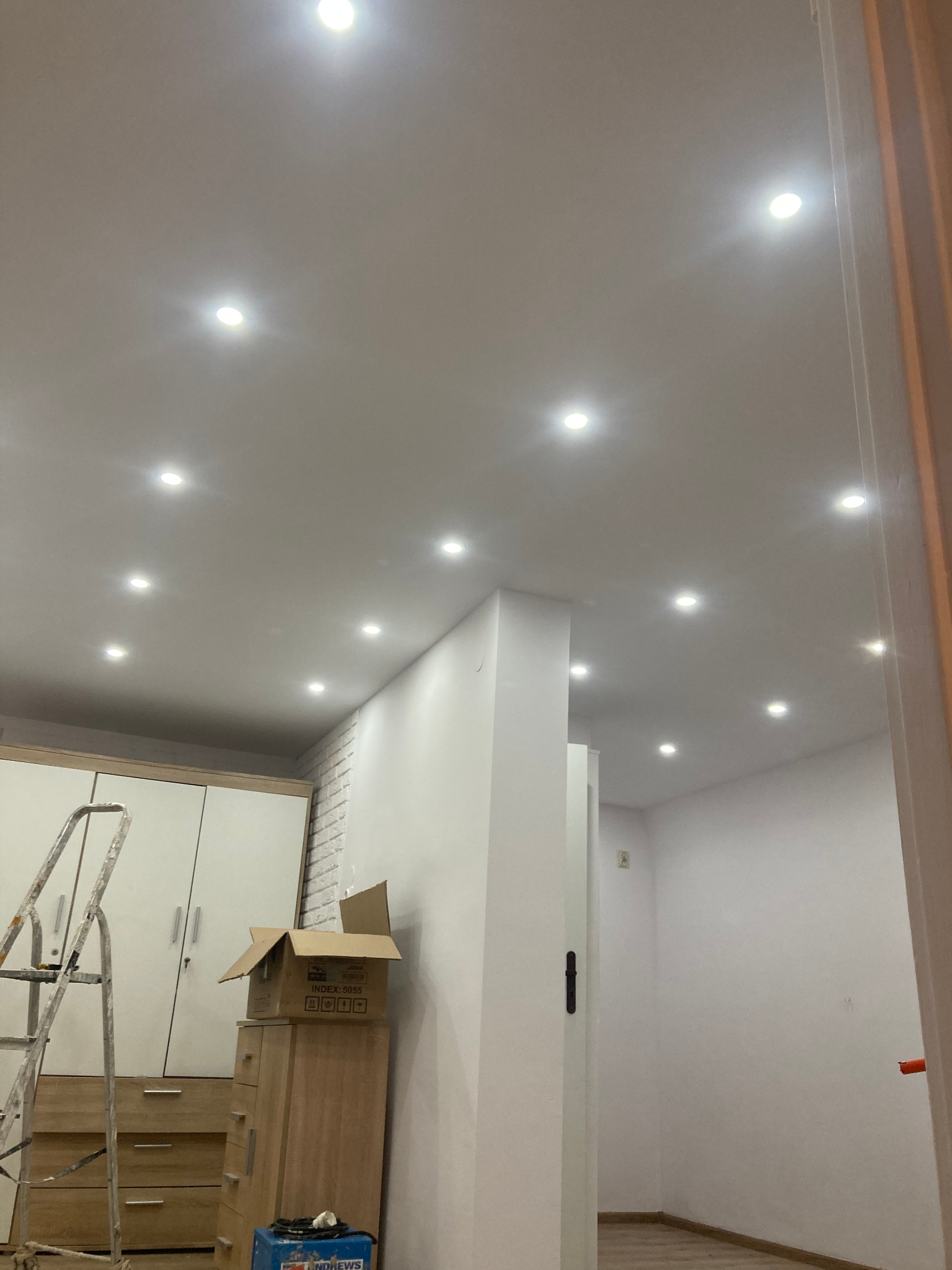 Jasne wnętrze po malowaniu z równomiernie rozmieszczonymi punktowymi lampami sufitowymi. Widoczny fragment drabiny i mebli w trakcie prac wykończeniowych.