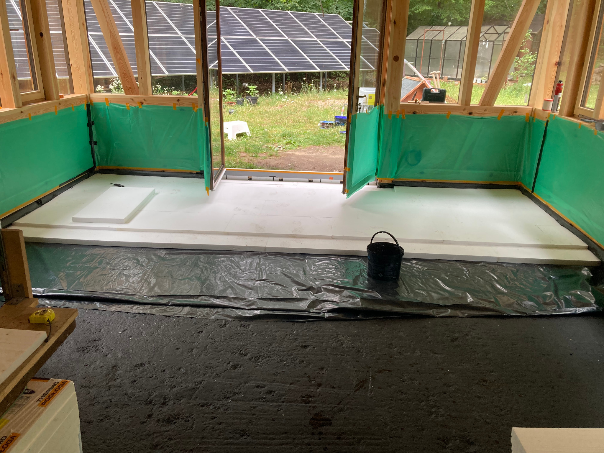 Ocieplanie podłogi w drewnianej konstrukcji z widokiem na panele słoneczne. Białe płyty izolacyjne na czarnej folii, zielona folia na ścianach. Prace wykończeniowe.