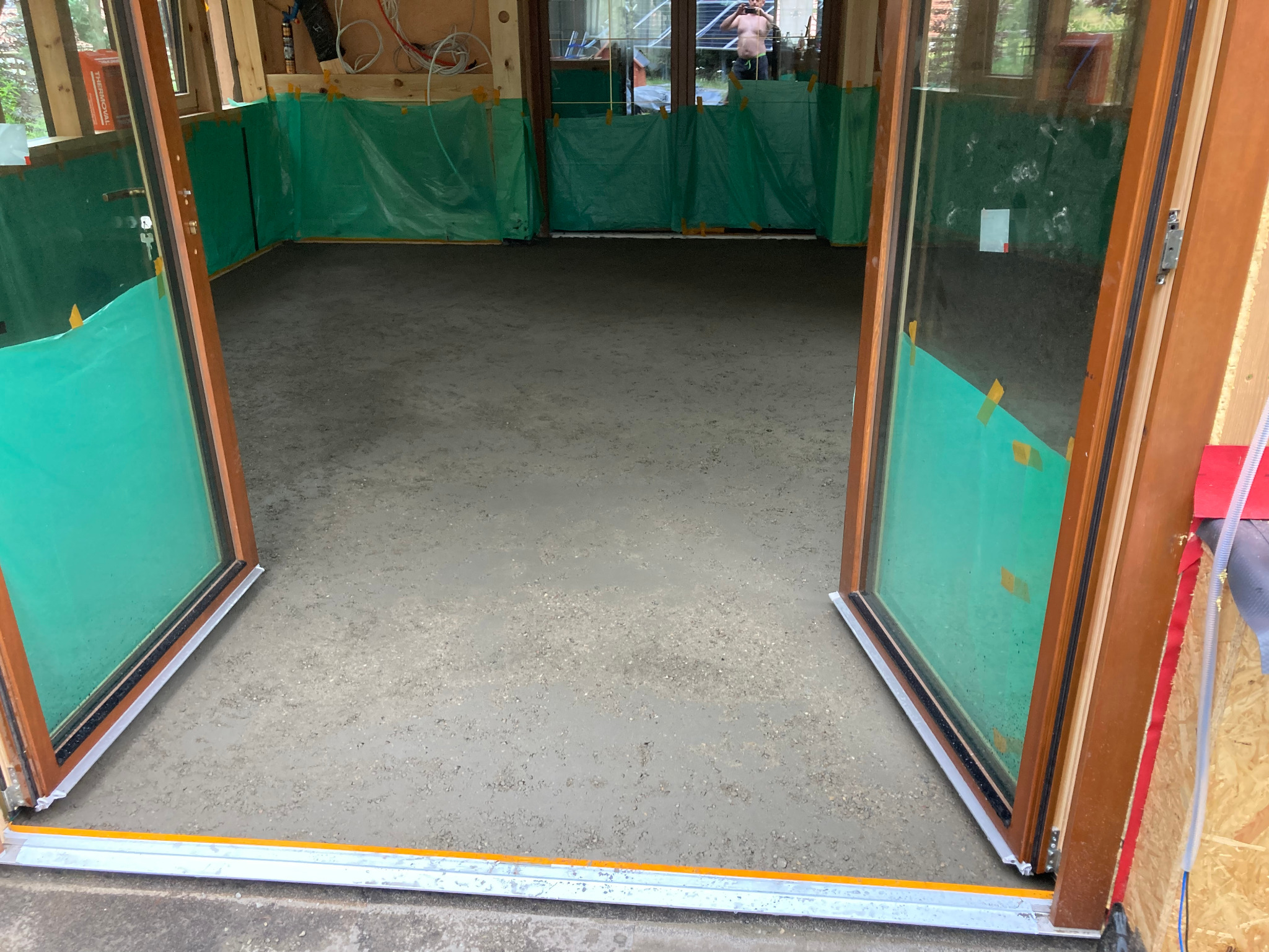 Wylana, świeża posadzka w pomieszczeniu z drewnianą konstrukcją i zabezpieczeniem folią. Widoczne otwarte, drewniane drzwi balkonowe z szybami.