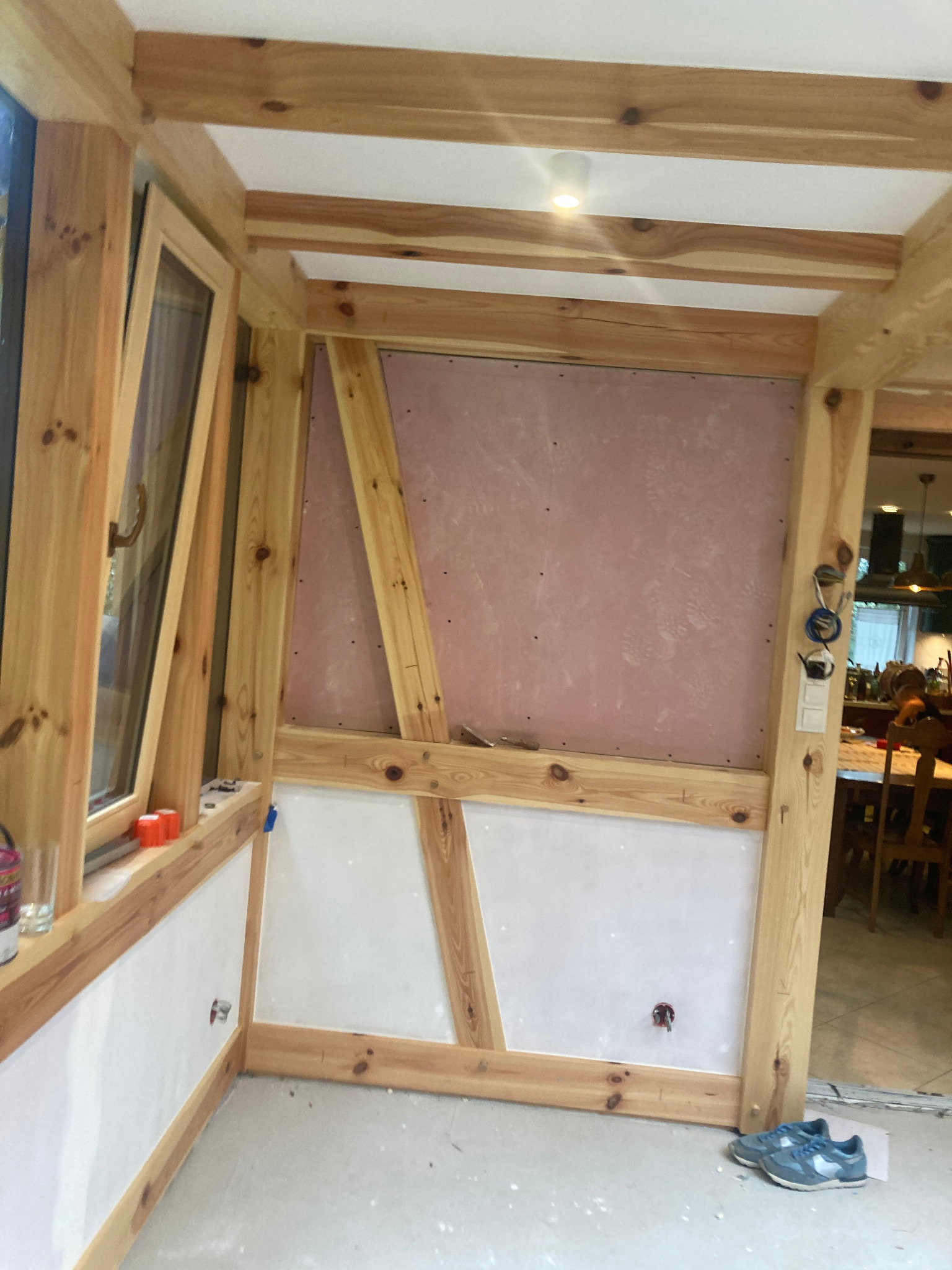Drewniana konstrukcja ścian z oknem, częściowo pokryta płytami. Widoczne instalacje elektryczne i para butów na podłodze. Prace wykończeniowe w trakcie realizacji.