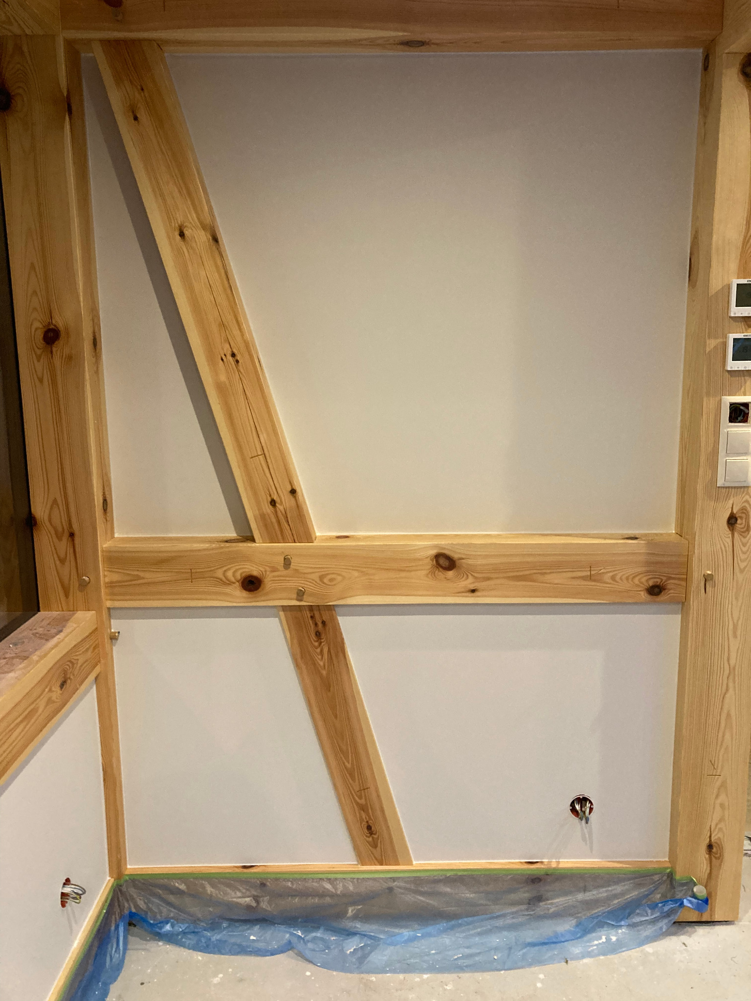 Ściana z drewnianą konstrukcją szkieletową, zabezpieczona folią malarską. Widoczne gniazdko elektryczne i włączniki. Świeżo pomalowana ściana w jasnym odcieniu.