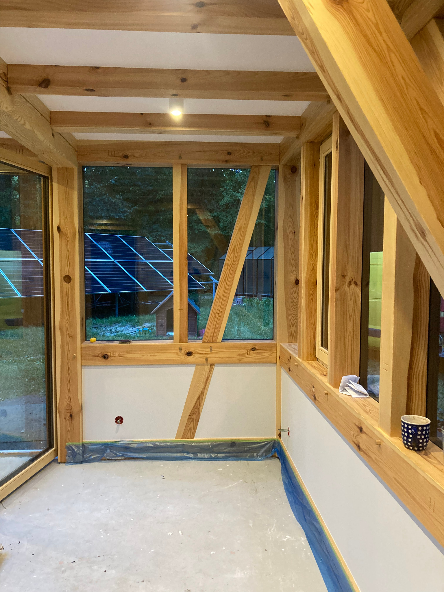 Wnętrze w trakcie wykańczania z drewnianą konstrukcją szkieletową, widoczne okna z panelami słonecznymi w tle, przygotowane do dalszych prac wykończeniowych.